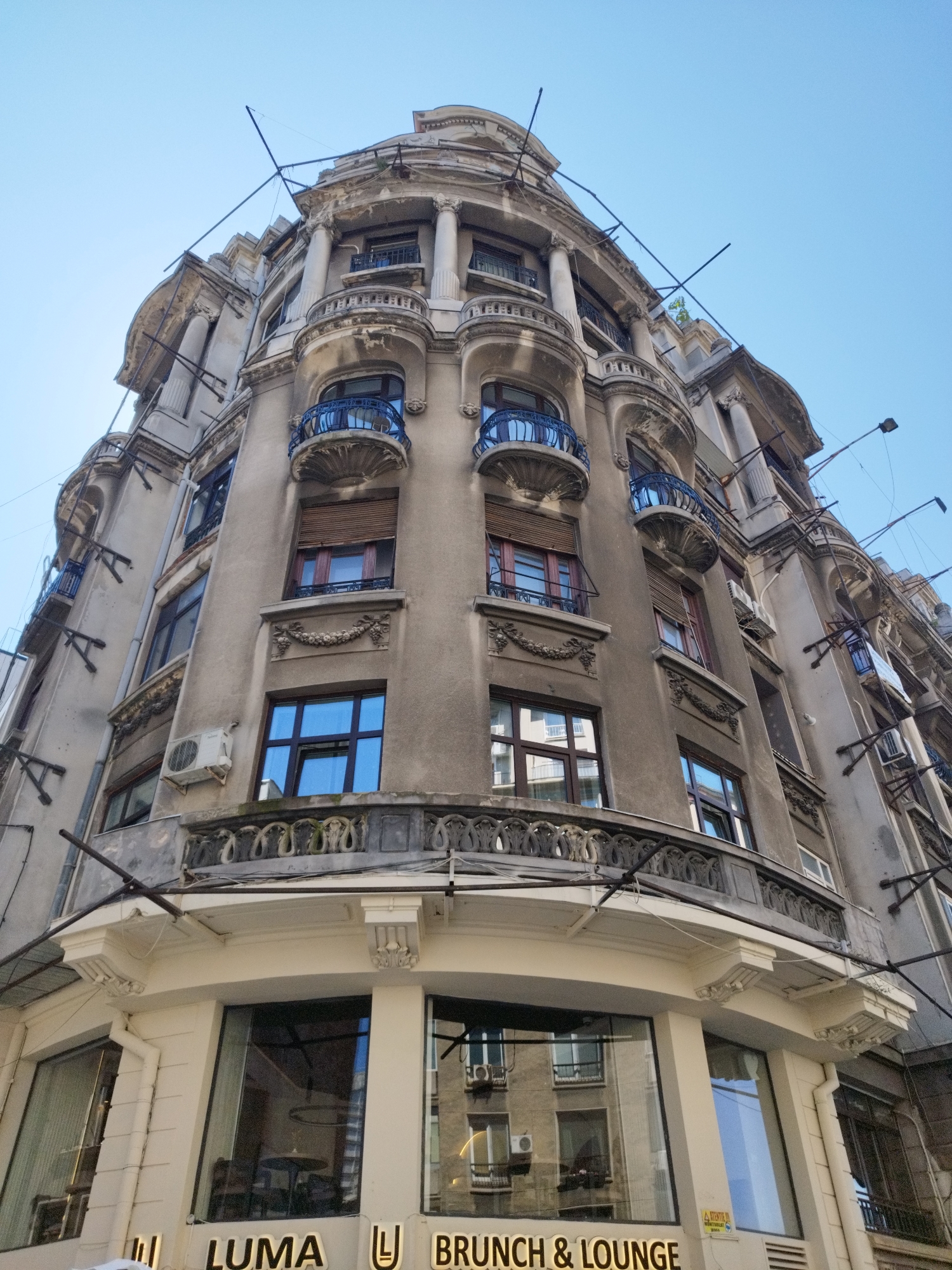 This low-angle, full-shot image captures the exterior of an old, ornate building in Bucharest, Romania. The building, likely a commercial establishment on the ground floor, exhibits classical architectural elements such as columns, detailed cornices, and wrought-iron balconies. The facade is primarily constructed of aged, greyish-brown stucco, with some sections appearing weathered and in need of repair. Several windows, varying in size and shape, are visible on the upper floors. Some feature dark blue wrought-iron railings, while others have wooden blinds. Air conditioning units are mounted on the walls.
The ground floor has large glass windows, reflecting the surrounding environment. Notably, illuminated signage for "LUMA" and "BRUNCH & LOUNGE" is visible, identifying the establishment. The sky above is a clear, pale blue. The overall impression is one of historical architecture in an urban setting, possibly showing signs of age and a blend of residential and commercial use. The presence of the signage confirms the location is in Bucharest, Romania.