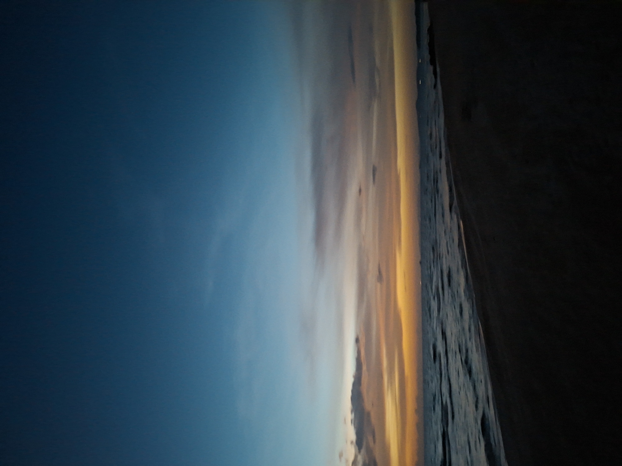 The image shows a beach scene at dusk. The sky transitions from a deep blue at the top to lighter shades of blue, orange, and yellow near the horizon, indicating the setting sun. Wispy clouds are scattered across the sky. The ocean is calm with small waves gently rolling onto the shore. The beach is dark and appears to be made of sand. There are no people visible in the image. The environment is an outdoor beach with the ocean and sky as the dominant features. The location is described as Unknown, Unknown.