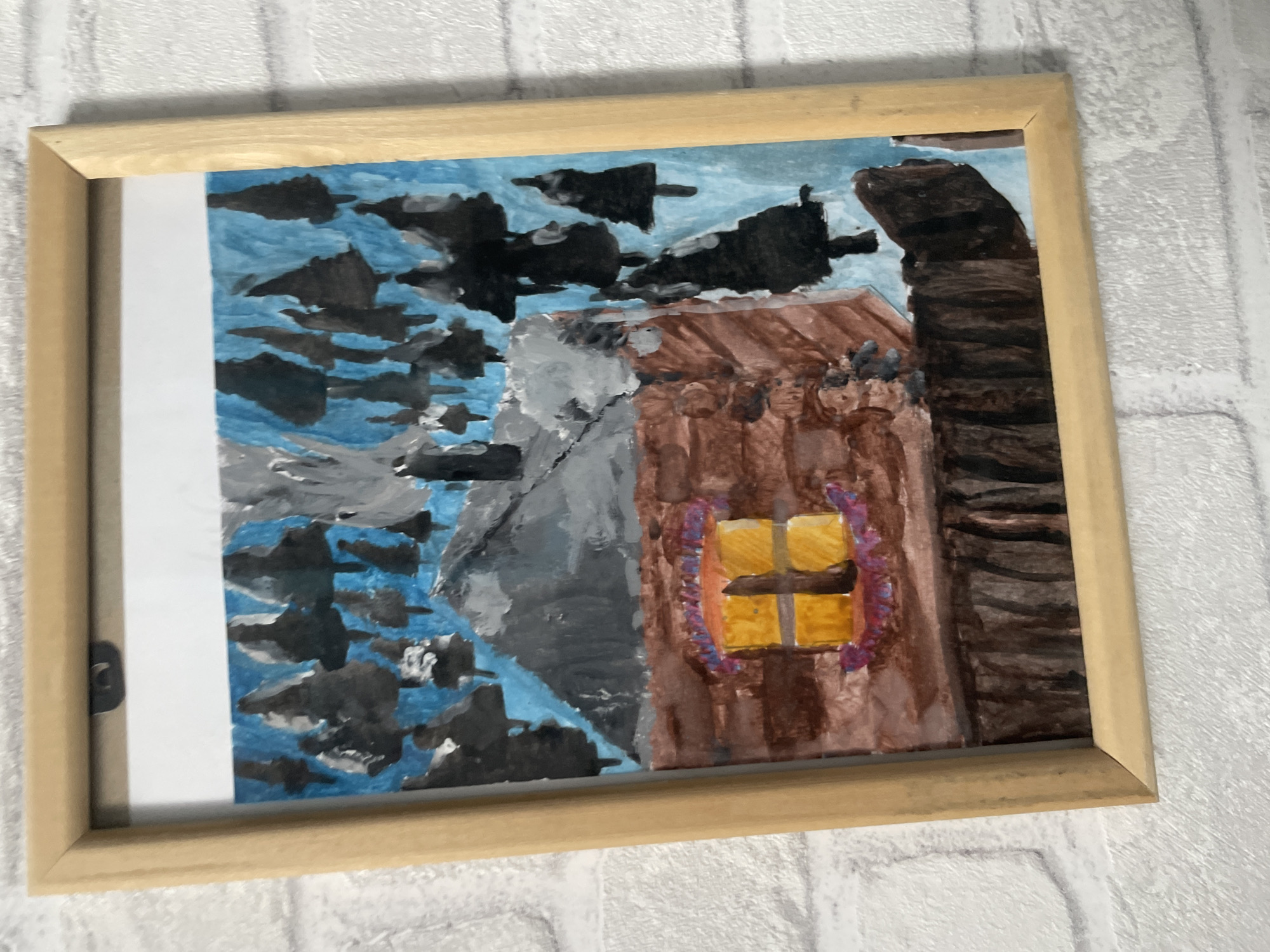 A framed watercolor or gouache painting is affixed to a white brick-patterned wall in the city of Chaikovsky, Russia. The artwork, oriented vertically, depicts a rustic log cabin under a blue sky populated by numerous dark evergreen trees, some with white accents suggesting snow. The cabin features reddish-brown walls with visible texture, a grey roof partially covered in white, and a dark grey chimney emitting lighter grey smoke. A prominent yellow-glowing window, bisected by dark vertical and horizontal lines, is centered on the cabin's front, surrounded at its base by purple and orange abstract shapes. The lower section of the painting shows a dark brown ground or foundation. The painting is encased in a simple light wood frame, which includes a metal hanger clip at the top center. The entire framed artwork is slightly tilted to the right.