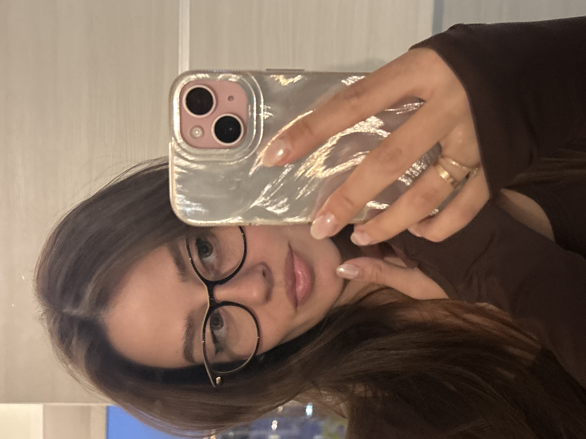 A young woman with brown hair and glasses is taking a selfie with her phone. She has light skin and is wearing a brown top. Her nails are painted with a clear coat. She is wearing rings on her fingers. The background appears to show a city skyline at night, suggesting she may be indoors near a window. Although the general location appears to be urban, it is not possible to verify if the photo was taken in Krasnoyarsk, Russia.