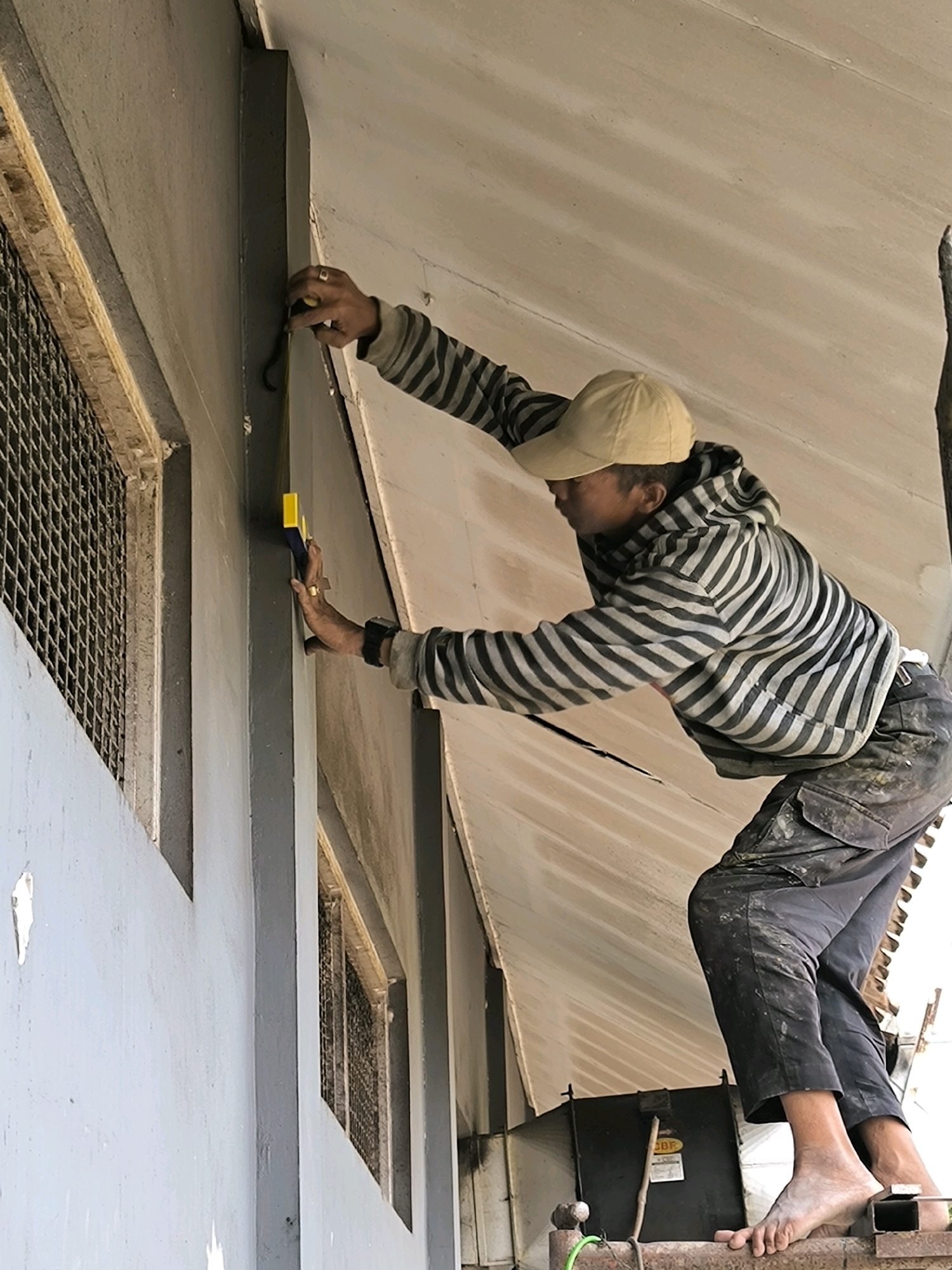 A person wearing a striped hoodie and a beige baseball cap stands on a metal structure. They are holding a tape measure against a grey painted wall and appear to be taking measurements. The person is barefoot and wearing dark pants stained with paint or dirt. In the background, a corrugated metal roof slopes upwards. The scene suggests an outdoor construction or renovation project in an urban setting.