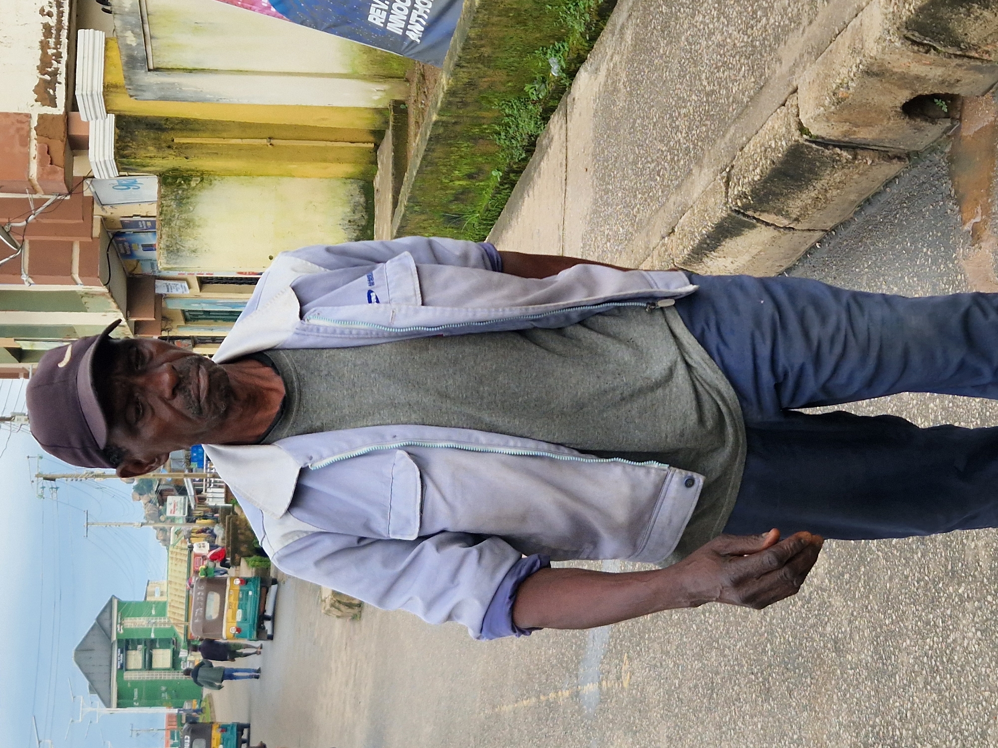 The image depicts a street scene in Kabong, Nigeria. In the foreground, an elderly Black man wearing a dark baseball cap, a light blue jacket over a grey t-shirt, and dark blue pants walks towards the camera. His right hand is in his pocket, and his left hand is visible, with fingers slightly splayed. The man has a weathered complexion and appears to be walking on a paved street.
In the background, the street is lined with buildings. To the left, a green, multi-story building with a prominent roof is visible. Further down the street, several small, colorful vehicles, resembling tuk-tuks, are parked or in motion. One of these vehicles, predominantly yellow and teal, has passengers visible inside. The buildings lining the street are varied in color and architectural style, with some appearing to be commercial establishments, judging by the signage and awnings. One building has a light yellow facade with green moss or algae growth on a low wall in front of it. Another structure, further to the right, features a large blue banner with white text. The sky is overcast. The overall environment suggests a busy urban or semi-urban area.