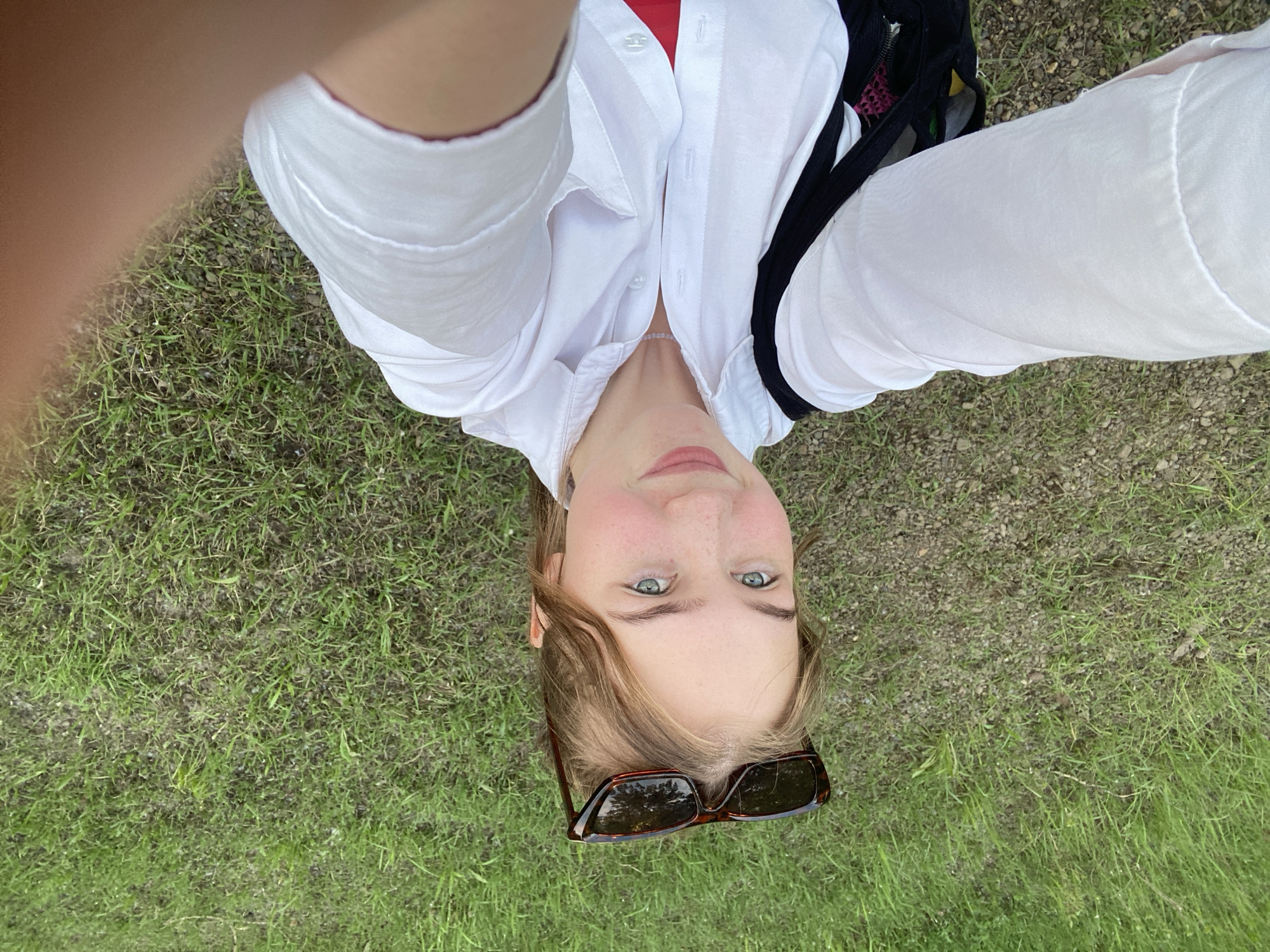 A young female subject is depicted in a selfie, outdoors in Chaikovsky, Russia. She has light brown hair, blue-green eyes, and a fair complexion with visible freckles across her nose and cheeks. A pair of tortoiseshell-patterned sunglasses with dark lenses is resting on top of her head. She is wearing a white long-sleeved button-down shirt, which is partially unbuttoned, revealing a red garment underneath. A light-colored beaded necklace is visible around her neck. A black strap, consistent with a backpack or shoulder bag, is visible over her left shoulder. The right hand, holding the camera, is partially visible in the lower right corner of the frame. The background consists of ground-level natural terrain, primarily composed of green grass interspersed with small grey and brown gravel and dirt.