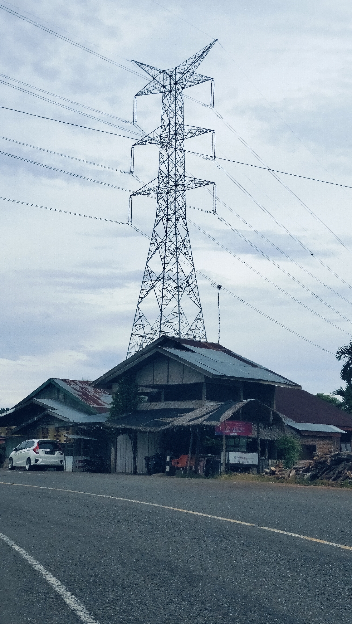 A vertical, eye-level shot captures a towering electricity transmission pylon against an overcast sky, with a cluster of buildings and a paved road in the foreground. The large lattice-style power tower, adorned with multiple power lines extending horizontally across the frame, dominates the upper and central portions of the image, silhouetted against the light grey, cloud-filled sky.
Below the massive pylon, a collection of rural or semi-rural structures line what appears to be a roadside. To the left, a white hatchback car is parked in front of a building with a corrugated metal roof. Adjacent to it, a central building features a steep, dark roof and an open-fronted section covered by a thatched canopy, under which chairs and an indistinct sign are visible; the sign appears to say "OPEN" and potentially some Indonesian text like "24 JAM". To the right, another building with a reddish roof stands, with a stack of firewood or logs nearby.
The foreground consists of a paved asphalt road, featuring a solid white line along its left edge and a solid yellow line marking the center. The diffused, natural light indicates daytime under a heavily cloudy sky. No individuals are present in the frame. The scene portrays a roadside environment in a residential or commercial area, consistent with the Lambarotunong, Indonesia context.