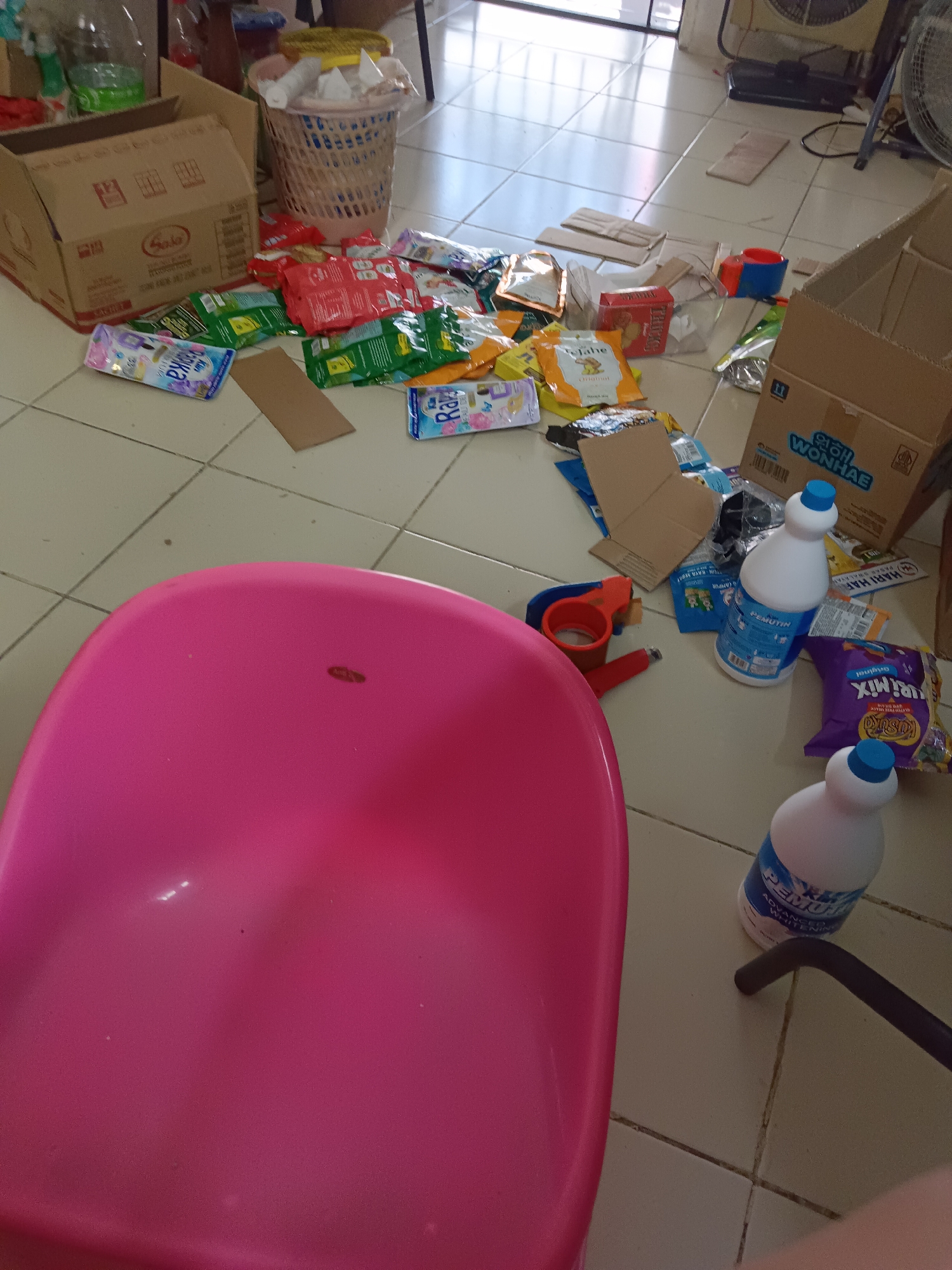 The image depicts a domestic scene of a cluttered floor in what appears to be a residential area. Several items are scattered across a tiled floor, including cardboard boxes, plastic packaging, bottles of cleaning supplies, and a pink plastic tub. A laundry basket filled with items is also visible. The lighting suggests an indoor setting, possibly a home or storage area. The specific location of Jayamukti, Indonesia is not discernible from the visual information present in the image. There are no individuals present in the frame, and no overt actions or interactions are being captured. The scene is static and focuses on the disarray of objects on the floor.