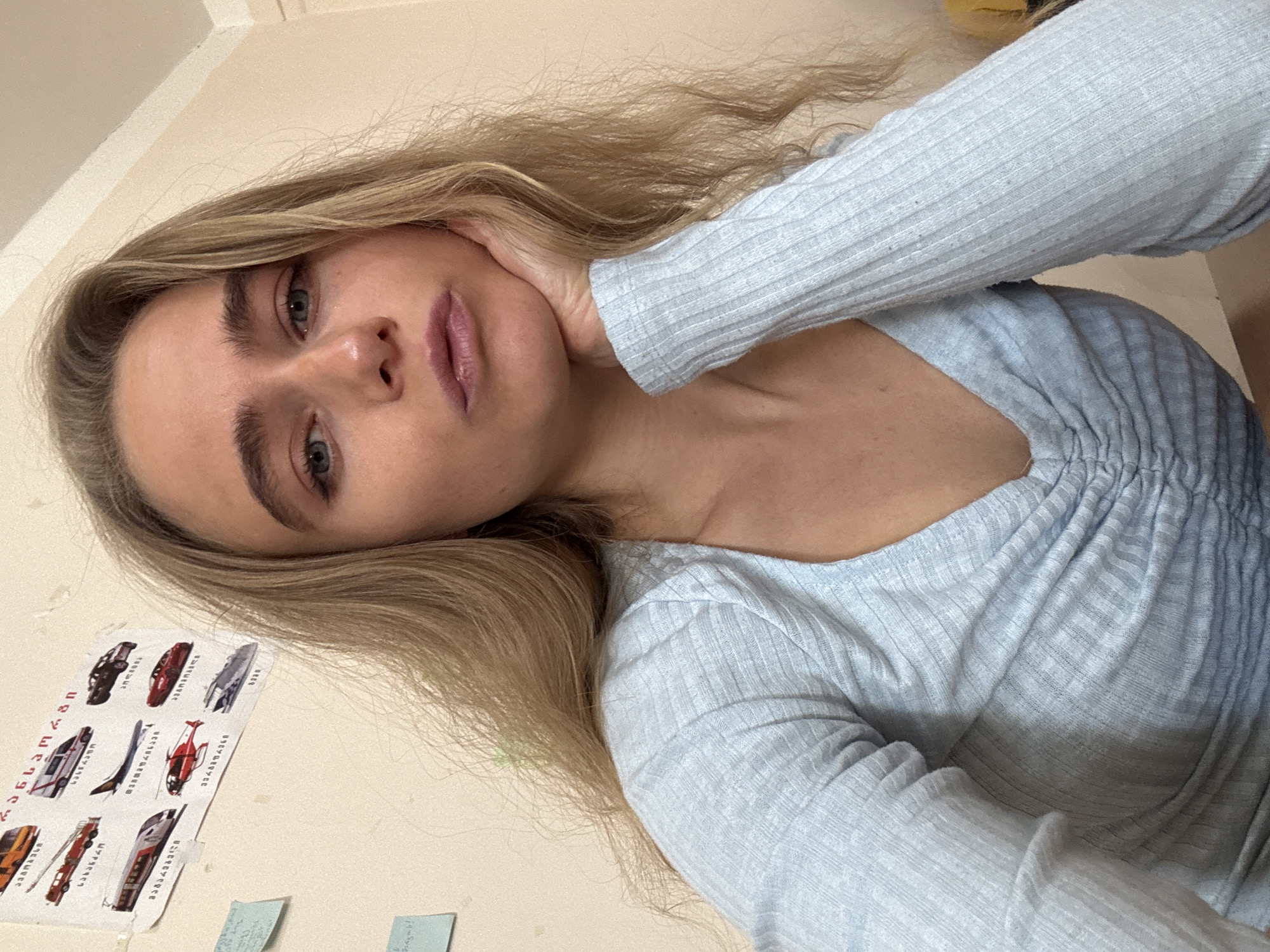 A close-up, tilted, indoor shot depicts a young woman with light blond hair, blue eyes, and full lips. She is wearing a light blue, long-sleeved, ribbed top. Her left hand is positioned against her face, resting on her cheek. The background consists of an off-white wall with a sticker sheet featuring illustrations of various vehicles and small, green sticky notes. The location is Unknown, Unknown.