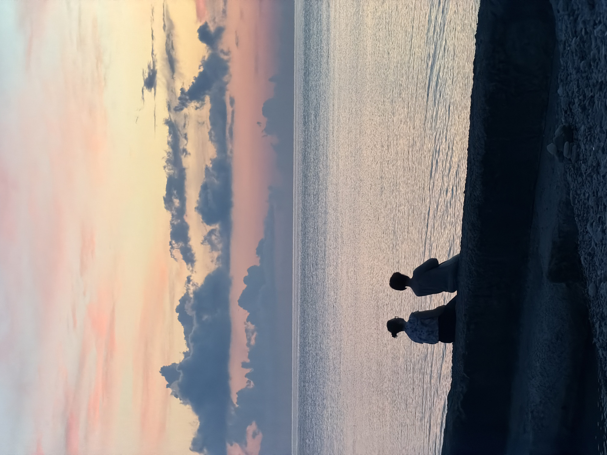 The image depicts a serene coastal scene at dusk, likely in Dederkoy, Russia, given the context. Two individuals are silhouetted against a tranquil sea, seated on a low, dark wall or embankment. The sky above is a gradient of soft hues, transitioning from a pale yellow at the horizon to pastel pinks and blues higher up, punctuated by scattered, textured clouds that are also tinged with pink and gray. The sea stretches out before them, its surface rippled and reflecting the colors of the sky, creating a sense of calm and vastness. The foreground is darker, showing the pebbled shore and the rough texture of the wall. The overall impression is one of quiet contemplation and the beauty of a fading day.