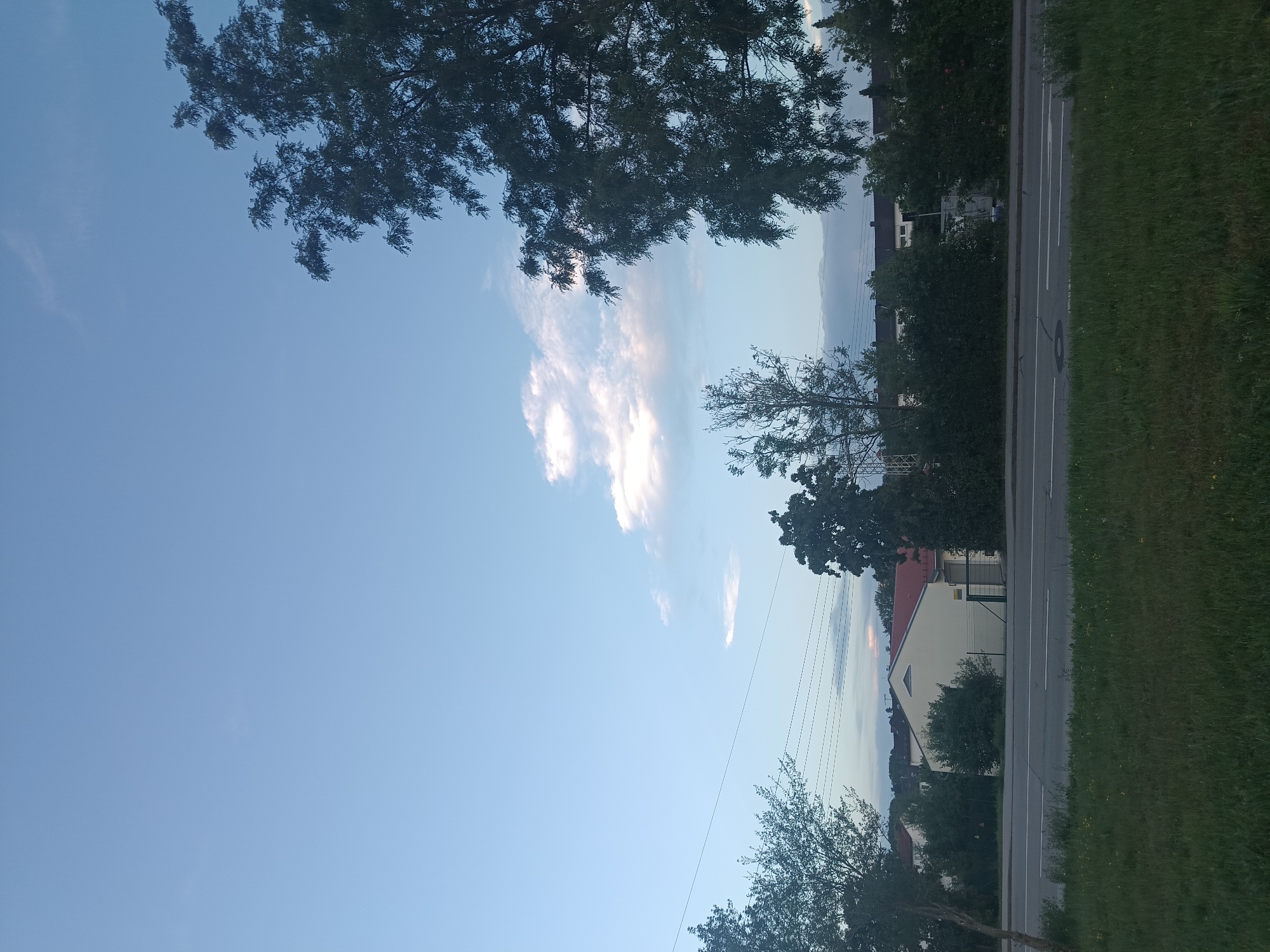 The image shows a street scene in Neuötting, Germany. In the foreground, there is a grassy area with a road running horizontally across the frame. The road has white lane markings. Behind the road, there is a row of trees and some buildings. The buildings appear to be residential houses with red tile roofs. The sky is blue with a few scattered clouds. The overall impression is a quiet suburban street.