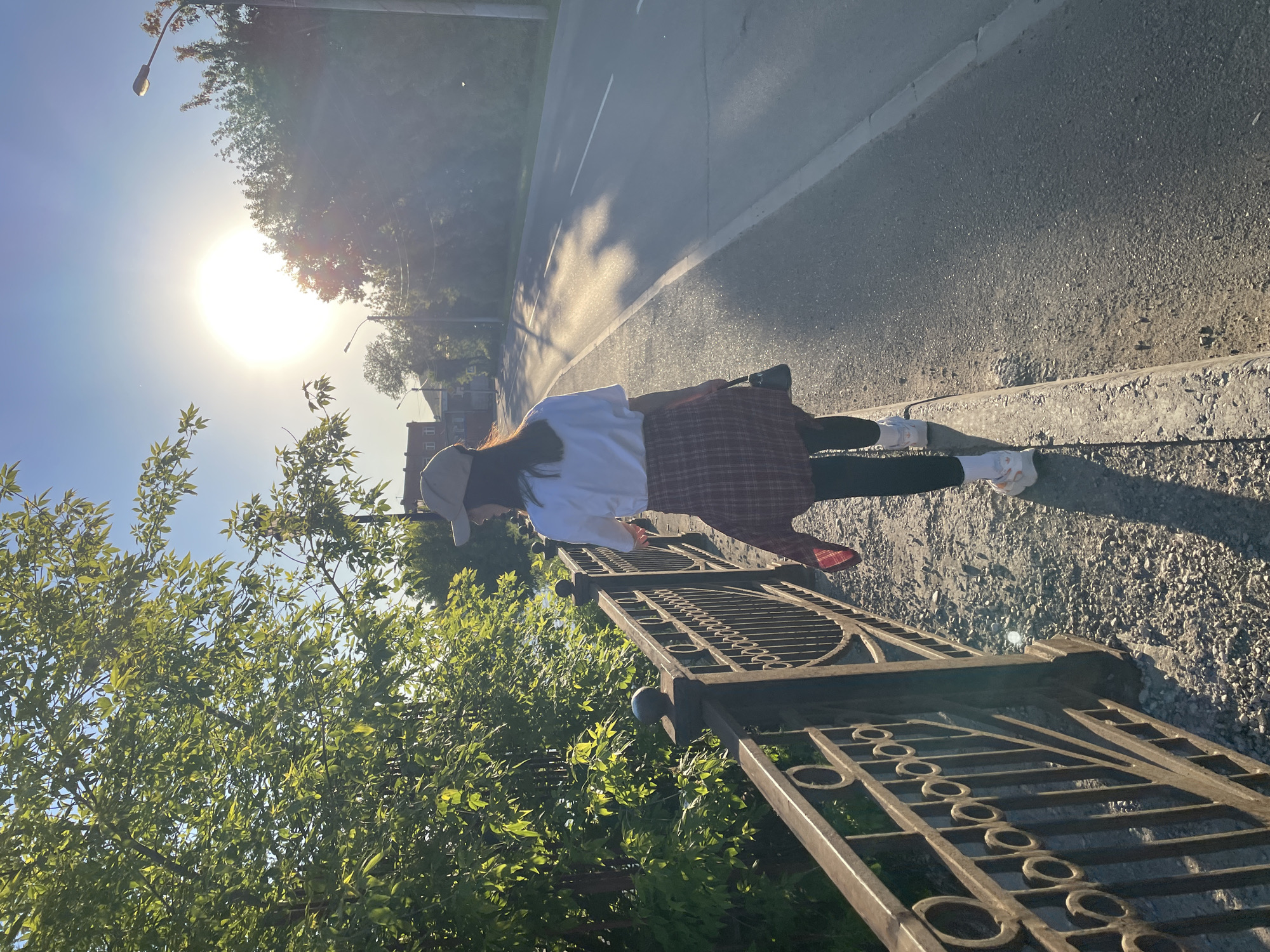 A young woman stands on a city street near an ornate metal fence in Krasnoyarsk, Russia. She is wearing a white t-shirt, a plaid skirt, black leggings, and white sneakers. A beige cap is on her head. The sun is high in the sky, casting long shadows on the pavement. A road runs parallel to the fence. Trees with green leaves are visible on the left side of the image, partially obscuring the sun.