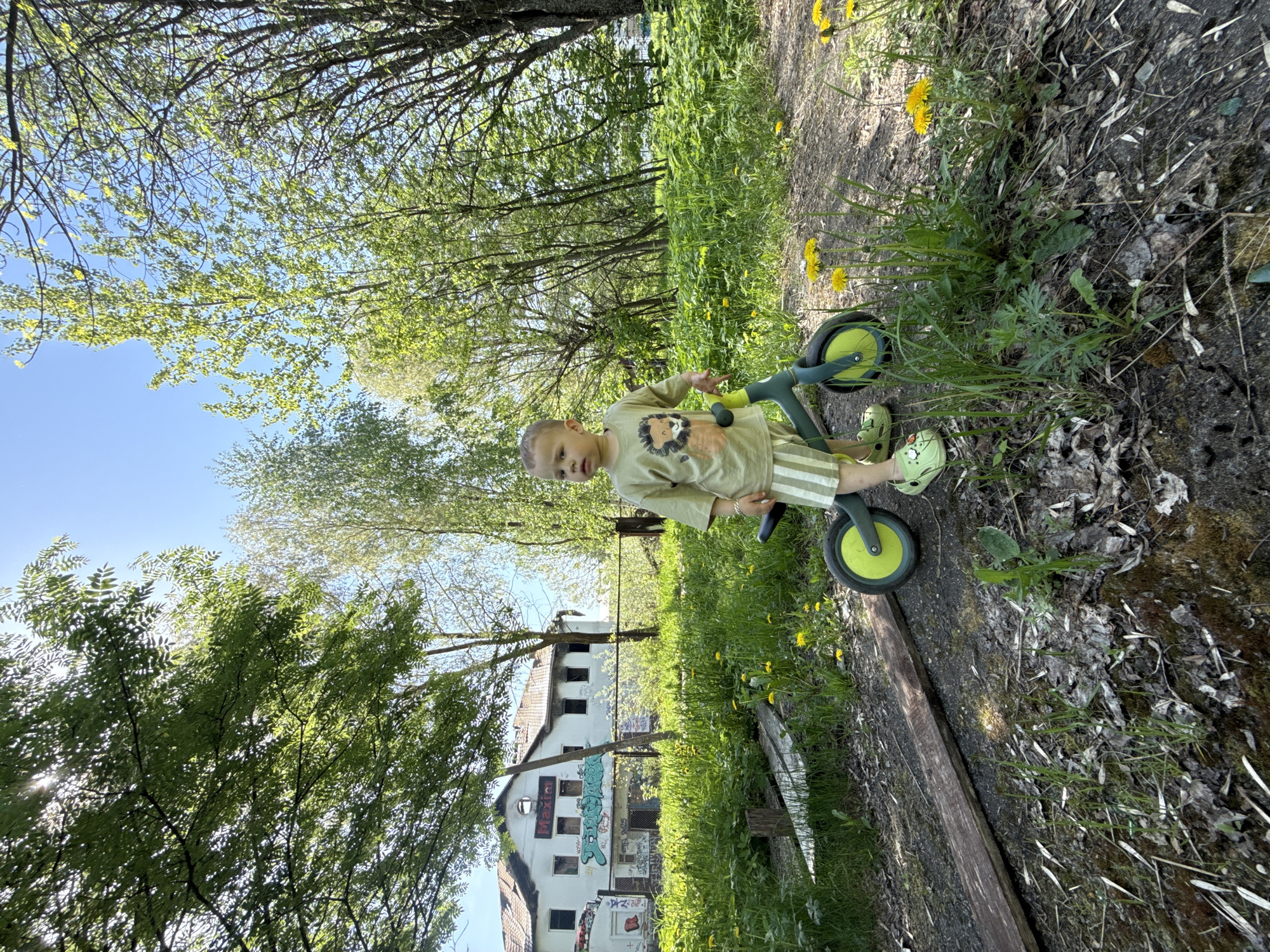 A young child stands next to a green balance bike on a grassy path. The child is wearing a green shirt with a lion graphic, striped shorts, and green clogs. Dandelions are scattered in the grass around the path. Behind the child and the path is a dilapidated two-story building with graffiti on its walls, partially obscured by trees and vegetation. The scene is located in Rusanovo, Russia, and the weather is sunny.