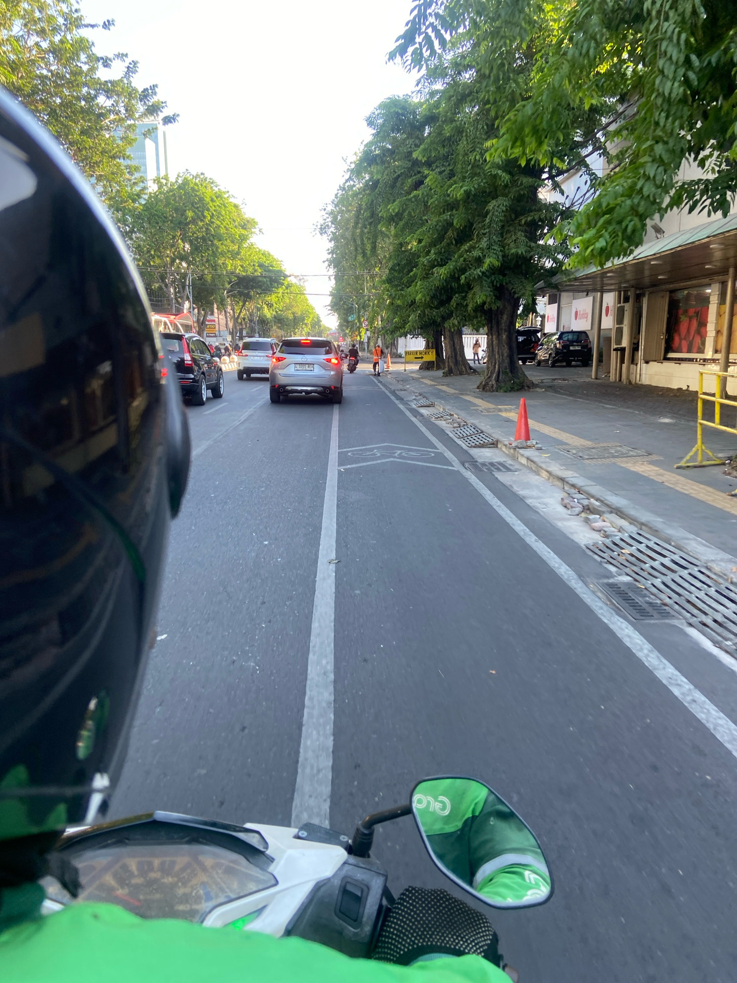 The image captures a first-person perspective from a motorcycle in Surabaya, Indonesia, on a street lined with trees and buildings. The rider, wearing a green jacket with a "GoFood" logo reflected in the motorcycle's mirror, is in motion, as indicated by the blur of the road and the perspective. Several cars are visible on the road, suggesting moderate traffic. On the right side of the road, a sidewalk runs alongside buildings, some with storefronts displaying products like strawberries. Orange traffic cones are placed along the sidewalk, and a yellow barrier is visible further down. The sky is bright, indicating daylight, possibly late afternoon or early morning, given the angle of the light. The overall scene depicts a typical urban environment in Surabaya during a period of activity.