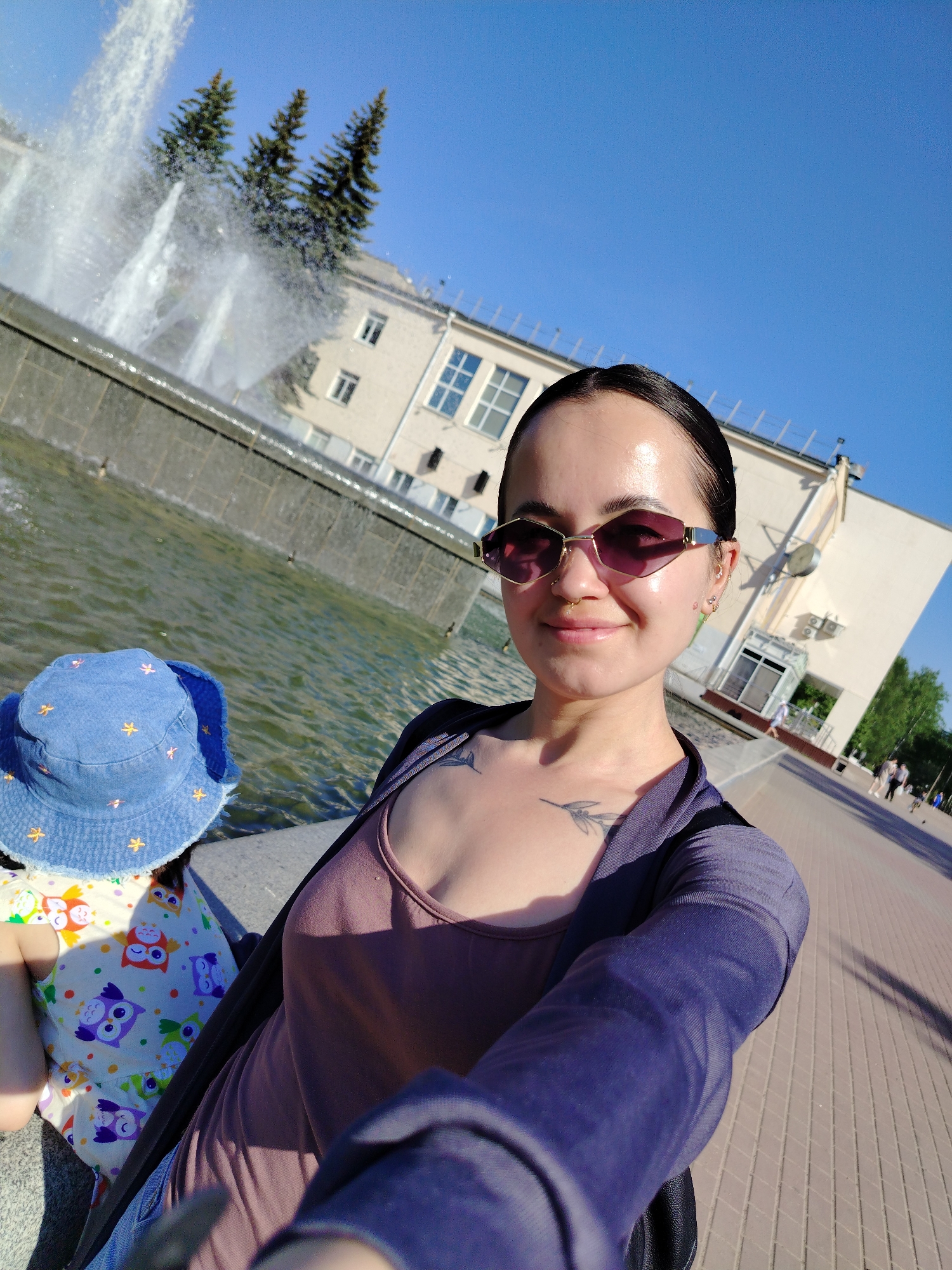 A medium, eye-level shot presents a woman taking a selfie beside a fountain and a child in the city of Chaikovsky, Russia. The woman, with slicked-back dark hair, wears angular purple sunglasses and a mauve tank top under a sheer navy blue cardigan. A bird tattoo is visible on her chest. She stands to the right, partially obscuring a child wearing a blue bucket hat with yellow stars and a dress adorned with owl patterns. They are positioned in front of a fountain with multiple jets spraying water upwards. In the background stands a light-colored, multi-story building with several windows and a central entrance. The sky is a clear blue. A brick-paved walkway stretches out to the right, with blurred figures of people visible in the distance.