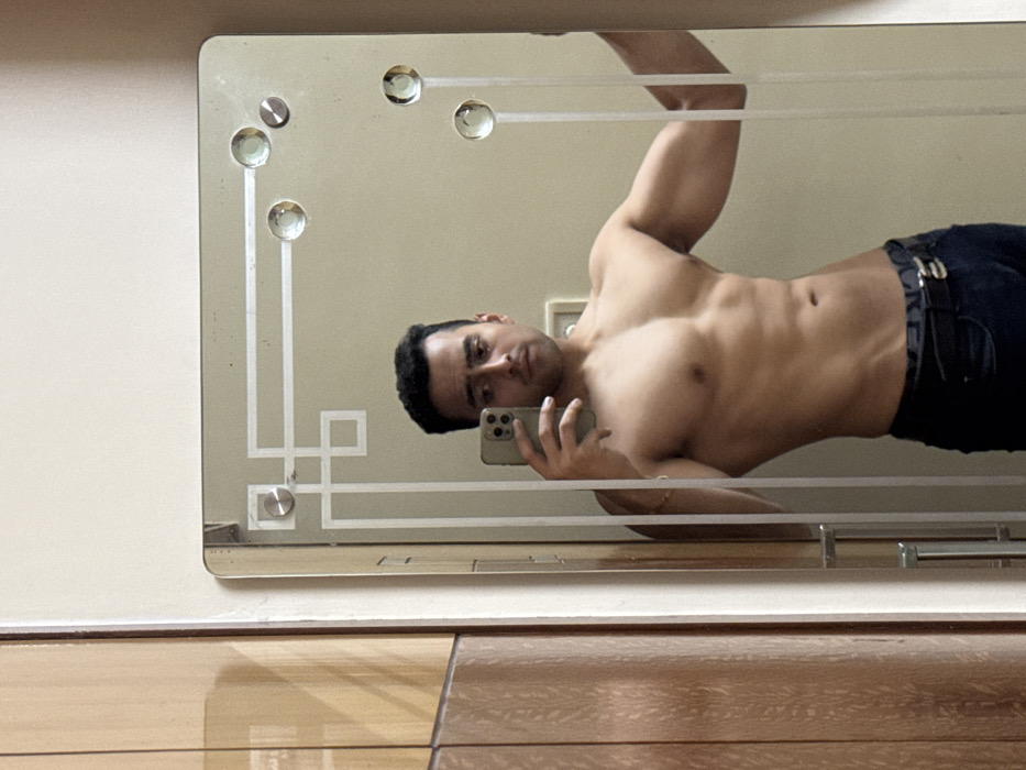 An indoor mirror selfie captures the reflection of a muscular man, believed to be in Kolkata, India, posing without a shirt. He is seen from the chest up, positioned diagonally within the mirror's frame. His left hand holds a smartphone, likely an iPhone with multiple cameras, to take the picture. The man has short, dark hair and a neutral to slightly serious expression, looking directly at his phone/reflection. He is shirtless, revealing a well-defined torso and arm muscles, and wears dark jeans or trousers with a dark belt and a metallic buckle.
The mirror itself is rectangular and features an ornate, etched border with geometric patterns, including lines and square shapes, and is adorned with several circular, gem-like or glass embellishments, along with metal fasteners. The mirror is mounted on a plain, light-colored wall. Below the mirror, a polished wooden floor is visible, reflecting some light. The lighting in the room appears even and bright, suggesting an indoor setting without specific time-of-day indicators. No discernible text is visible on any of the subjects or objects in the image.