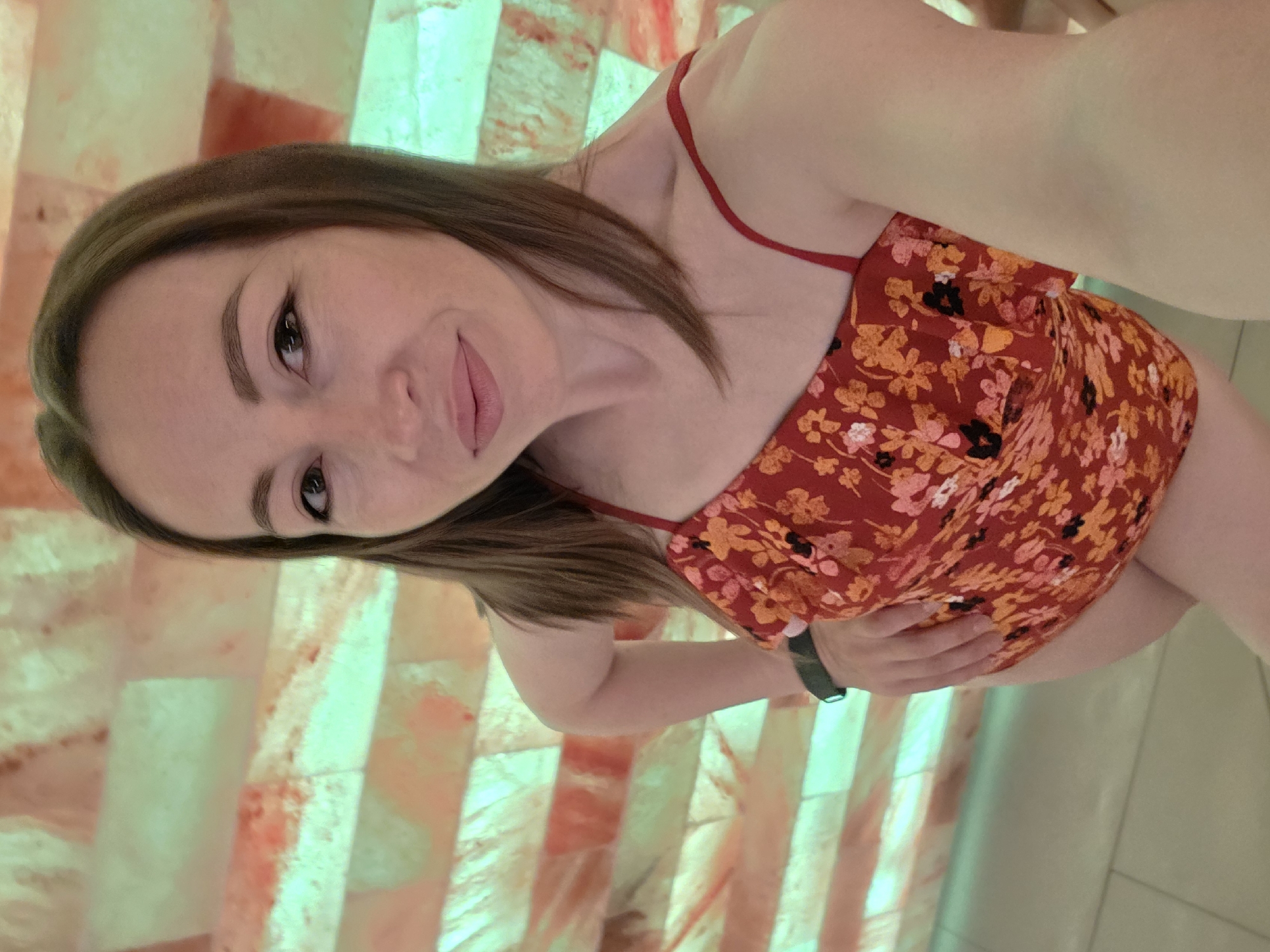 A woman is captured in a selfie, looking directly at the camera with a gentle, inviting smile. Her head is tilted, and her long, brown hair falls over her shoulders. She wears a red top adorned with an orange, pink, and black floral pattern, featuring thin straps. A dark-colored smartwatch is visible on her left wrist.
The background consists of an illuminated wall made from rectangular blocks that appear translucent, possibly salt blocks, given the common design of salt rooms. These blocks glow with varying shades of orange, pink, and a soft greenish-blue, creating a warm and diffused ambient light. The overall setting suggests an indoor environment like a spa or wellness center. The image is rotated, indicative of a handheld selfie. There is no visible text in the image. The location context provided for this image is Chaikovsky, Russia.