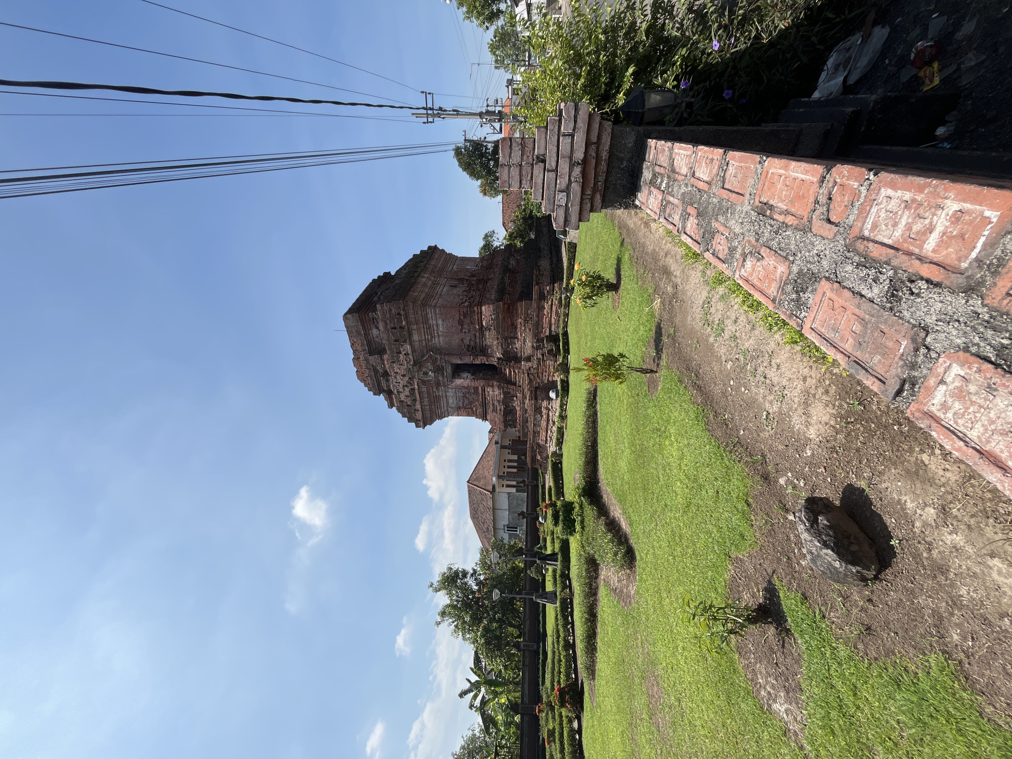This image shows a red brick temple structure in the city of Wunut, Indonesia, under a clear blue sky with scattered clouds. The temple is surrounded by a well-maintained green lawn and garden area, featuring manicured hedges, small trees, and pathways. A brick wall with decorative elements runs along the foreground, partially obscuring a lamppost and lush green foliage. In the background, there are houses and trees, with telephone wires and a utility pole visible on the right side of the frame. The scene is a sunny day, suggesting pleasant weather. There are no people visible in the image.