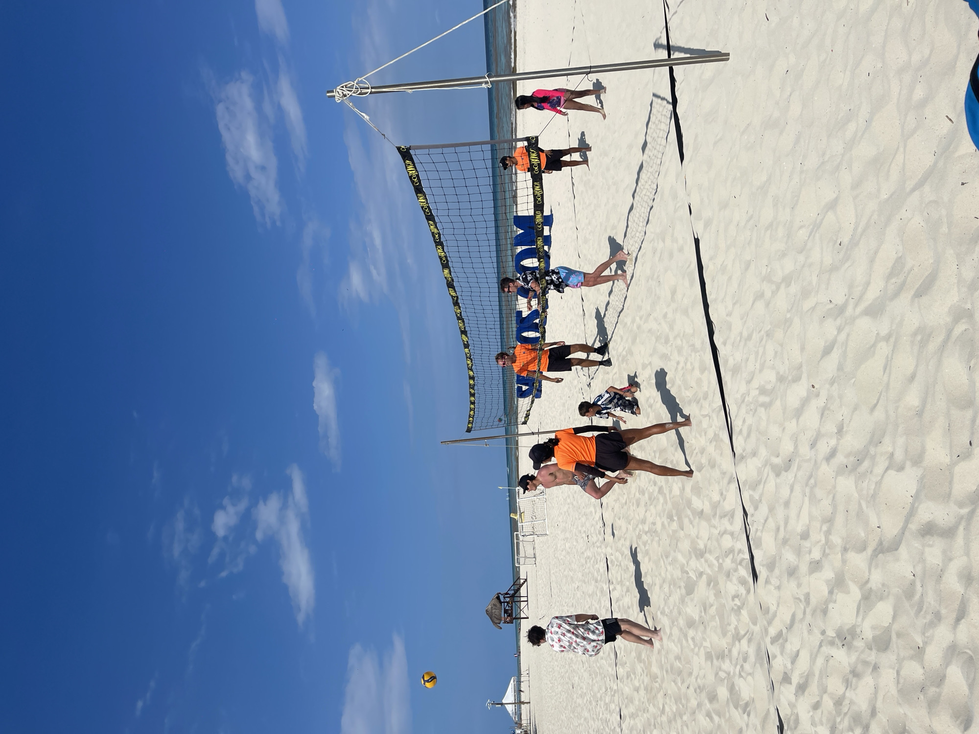 A group of people is playing beach volleyball at an unidentified beach in Playa del Carmen, Mexico. The volleyball net is set up on a white sand beach under a blue sky with scattered clouds. There are ten people in the scene, including seven players near the net, one player further away, and two individuals near the water's edge. The volleyball is visible in the lower-left part of the frame. The sun is shining brightly, casting shadows from the people and the volleyball net onto the sand. In the background, there are some beach structures like a small hut with a thatched roof and some white canopies.