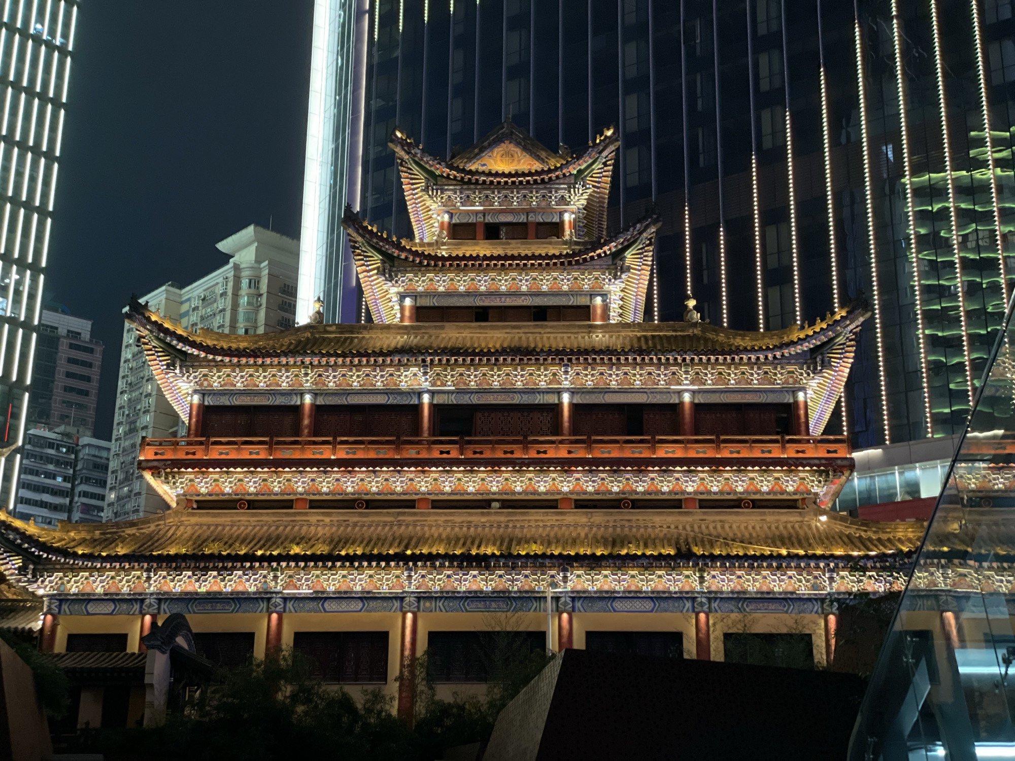 Captured at night, the photograph showcases a traditional Chinese building with multiple tiers, prominently lit with yellow and white lights. This structure, decorated with intricate details and patterns, is situated in Yuzhong District, China, a locale known for its blend of historical and modern architecture.
The traditional building is juxtaposed against a backdrop of modern skyscrapers, some of which are illuminated with vertical lines of white and yellow lights, indicating its location in a dense urban setting. The sky is dark, suggesting a nighttime scene.
Vegetation and architectural elements are visible in the foreground, creating depth and context for the main subject. The image captures a blend of traditional Chinese architecture amid a contemporary urban landscape, characteristic of the Yuzhong District.