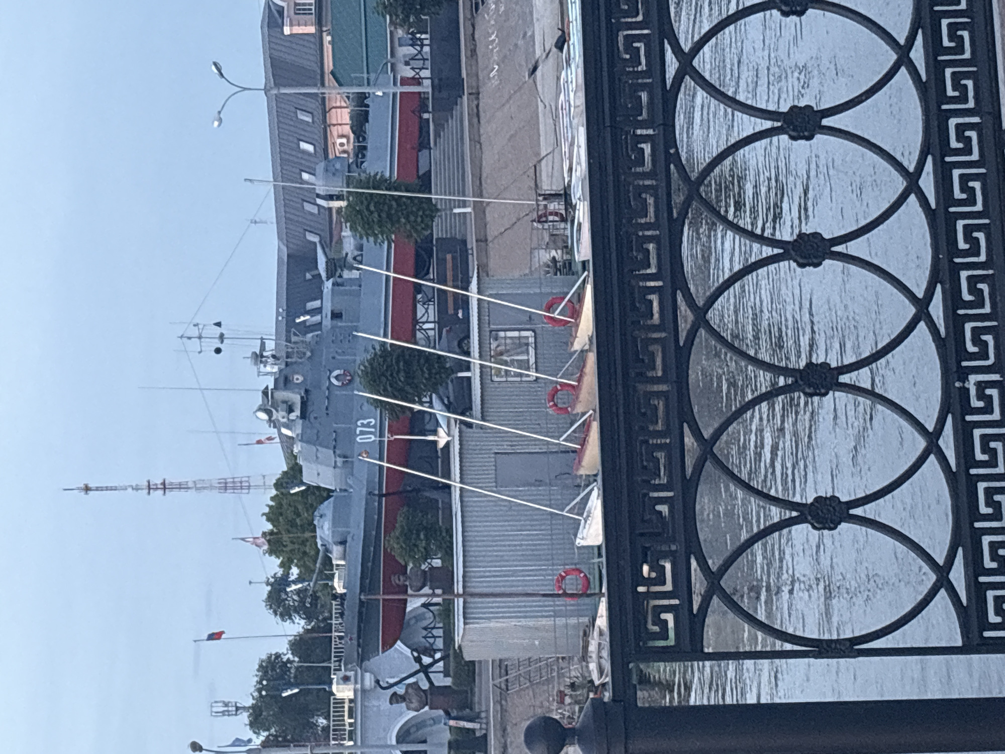 The image captures a view of the Astrakhan Kremlin's naval museum, specifically the "Salty Koster" (Salt Bonfire) cruiser. The main subject is the museum ship, identified by the number "073" on its hull. The cruiser is gray with a red stripe along its waterline. In front of the cruiser, a row of small boats, possibly rowing boats or kayaks, are lined up. Several lifebuoys are visible, some red and others white. Behind the boats, a corrugated metal building is present, with a ladder leaning against it. Two statues of sailors stand on the embankment near the ship. In the background, buildings with gray roofs and windows, a radio tower, and trees are discernible. The scene overlooks a body of water, with a decorative black metal fence in the foreground. The sky is overcast, suggesting it is either early morning or late evening.