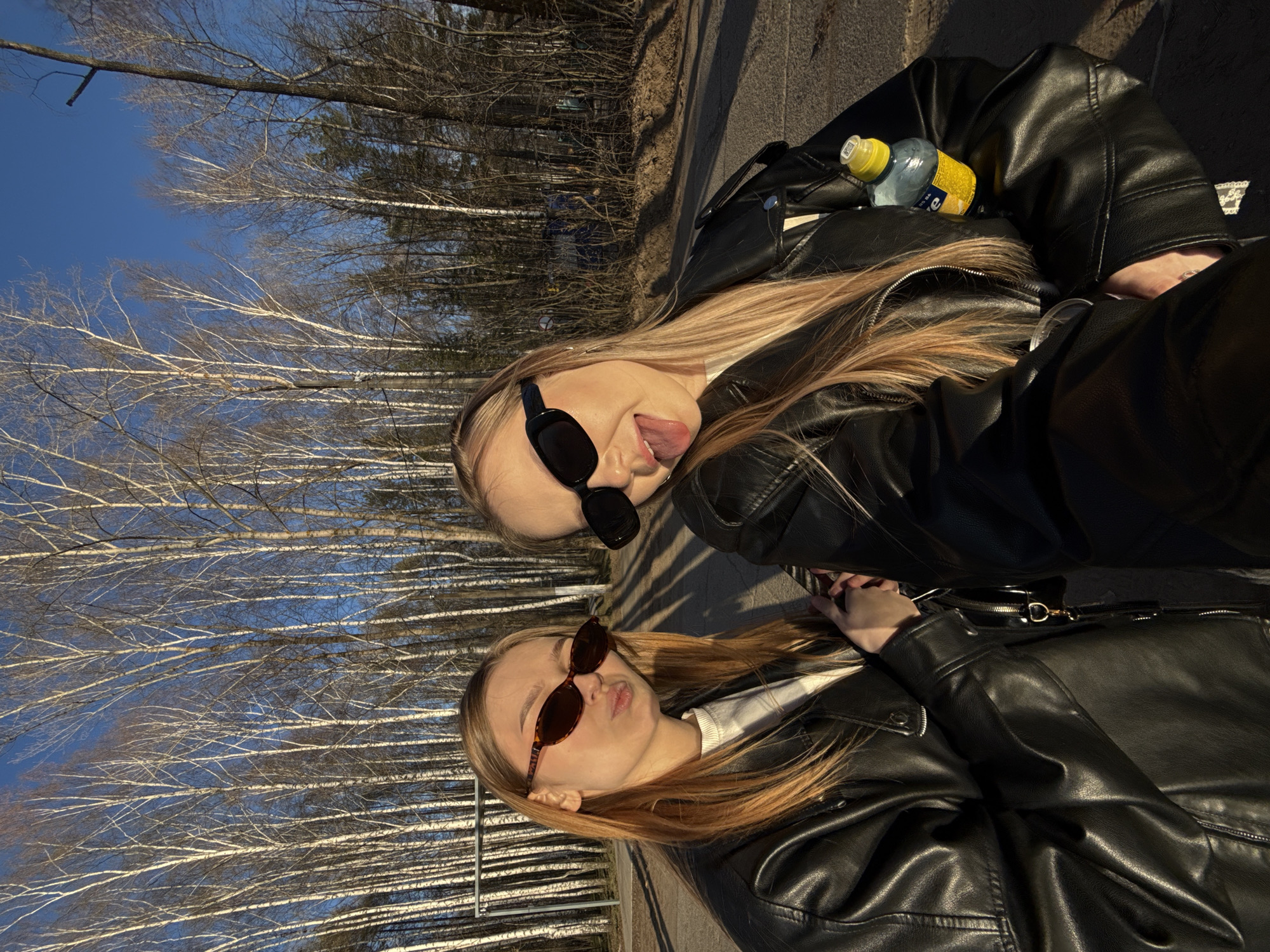 Two young women pose in an outdoor environment, likely in Chaikovsky, Russia. The upper woman is sticking her tongue out and is wearing dark sunglasses and a black leather jacket. A bottle of water is visible near her. The lower woman is also wearing sunglasses and a black leather jacket. The background features leafless trees against a blue sky and a paved surface.