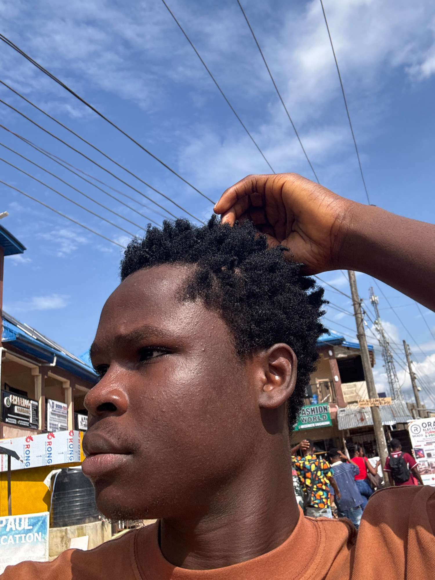 A young man is photographed from the side in Ihiagwa, Nigeria. He has dark skin and short, curly black hair, with a hand gently touching his head from behind. He is wearing an orange t-shirt. In the background, a street scene unfolds with buildings, storefronts displaying signs, including one that reads "FASHION WORLD," and utility poles with a network of electrical wires overhead. Several people are visible walking in the background, and a cell tower stands in the distance. The sky is blue with scattered clouds.