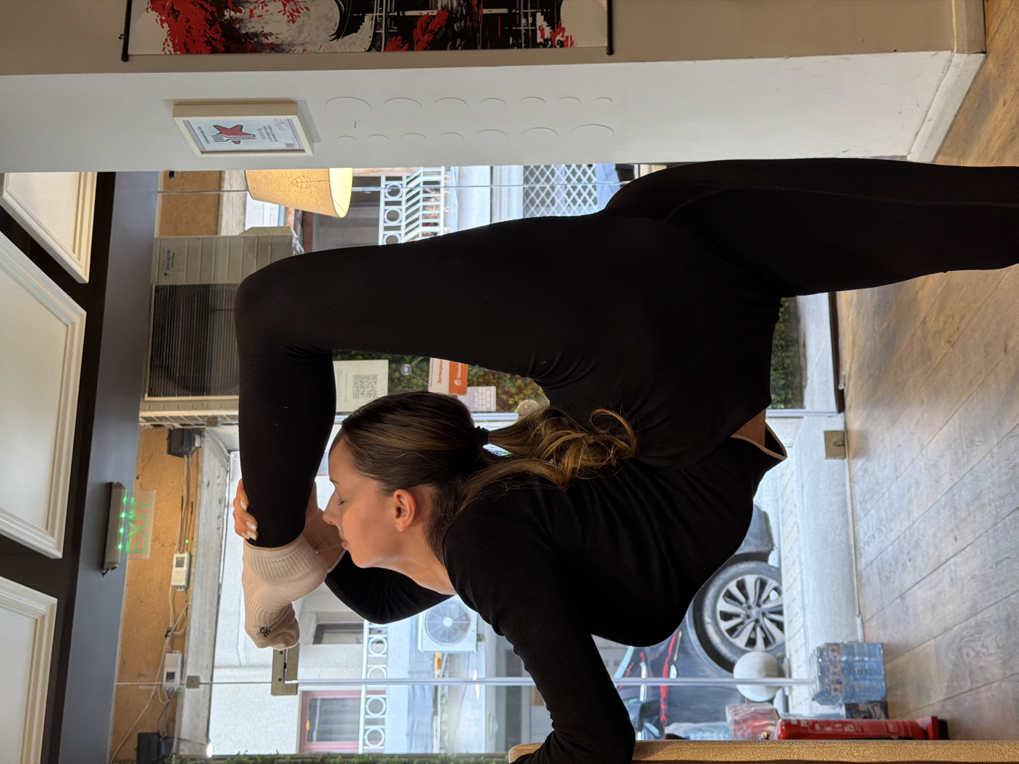 A woman in black athletic wear is performing a complex yoga pose indoors. She is balancing on her hands on a wooden surface. Her head is bent forward, with one of her feet touching her face. She is wearing a light-colored sock on that foot. Her hair is pulled back in a ponytail.
The background shows an urban environment, visible through a glass window. The exterior view includes buildings with multiple windows and air conditioning units, a car, and various signs and objects on the street.
Based on visual cues like architectural styles, signage, and the general ambiance of the surroundings, the location is consistent with a city like Sofia, Bulgaria, where older European architectural styles and modern elements often coexist. The specific street or neighborhood cannot be pinpointed without further information.