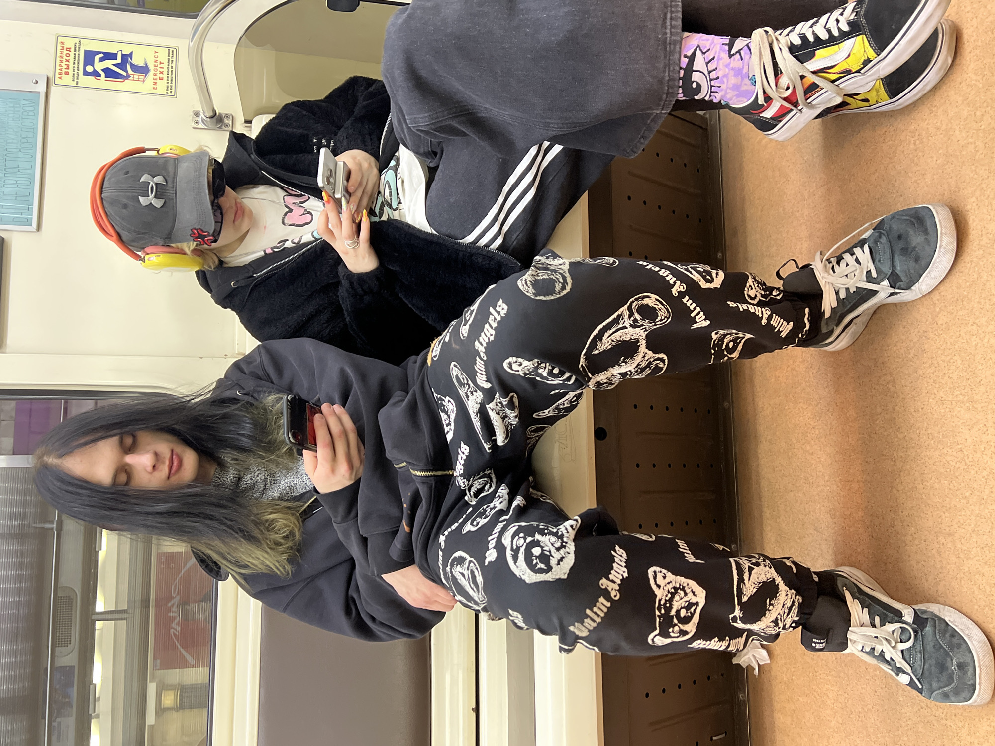 Two people are sitting next to each other on a bench seat inside a subway train in what appears to be Shushary, Russia. The person in the foreground has long, dark hair with streaks of blonde, is wearing a black hoodie and black pants with white bear designs and the words "Palm Angels" on them, and is holding a phone. They are also wearing dark-colored sneakers. The person in the background is wearing an orange headset, a gray cap, sunglasses, a black jacket, white shirt, and black Adidas track pants. They are also holding a phone. They are wearing colorful socks with cartoon designs and brightly colored sneakers. The interior of the train is visible, with beige walls and seating. An emergency exit sign is visible in the background.