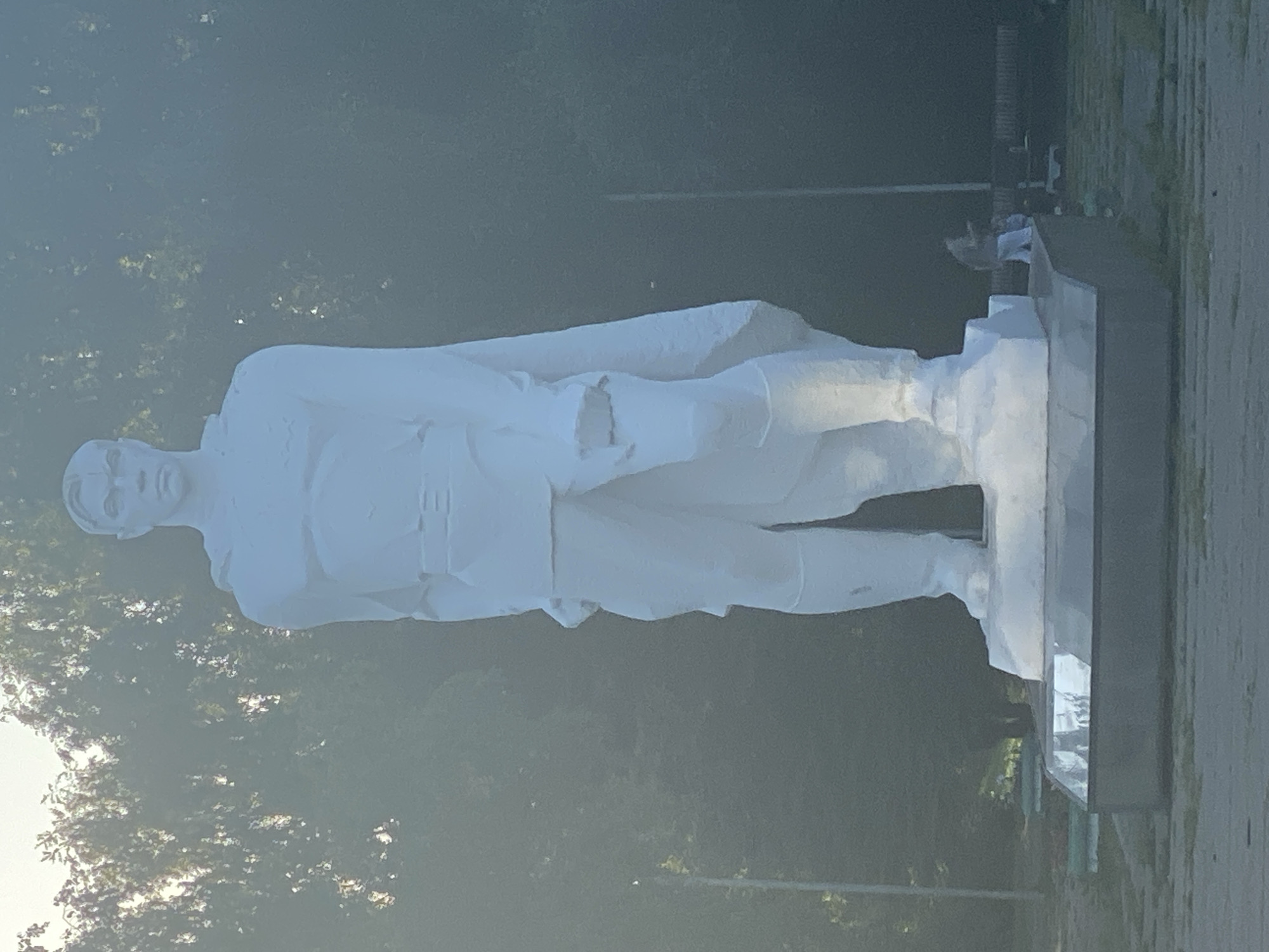 A large, white, full-body statue of a man stands centrally in the image. The statue depicts a figure with a stern expression, wearing a uniform with a cape draped over one shoulder. The man is standing with his hands at his sides. The statue is mounted on a multi-tiered, grey base, which rests on a reflective, polished surface. In the background, trees with lush green foliage are visible, creating a soft, hazy backdrop. The sky is a pale, bright color, suggesting it is either early morning or late afternoon. To the right of the statue, two figures are faintly visible in the distance, appearing to be walking or standing near benches. The ground in front of the statue is paved. The overall scene appears to be an outdoor public space, possibly a park or memorial, located in Novokuznetsk, Russia.