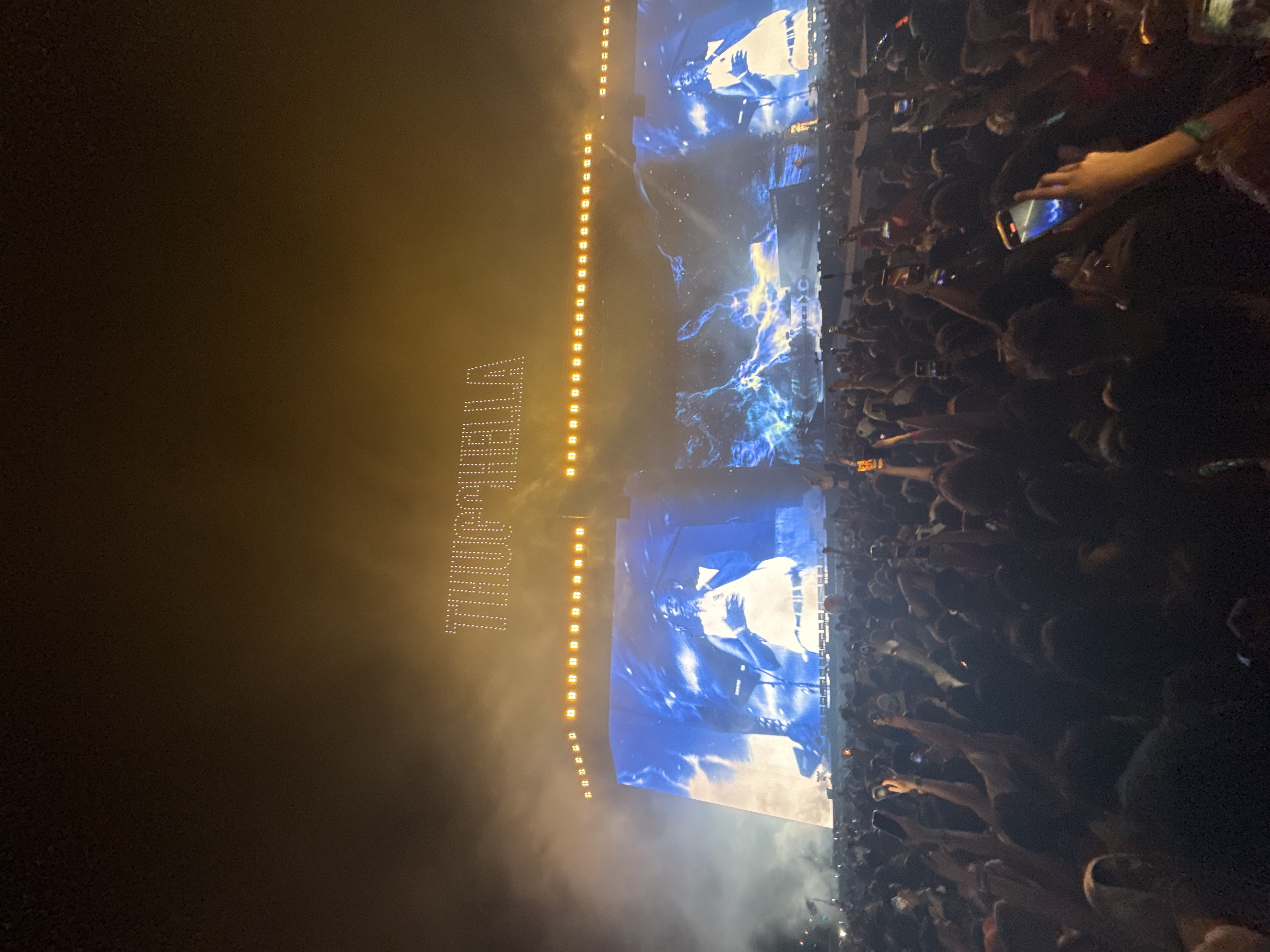 A large crowd of people is gathered at night in Indio, California, United States, attending what appears to be a concert. Many individuals are holding up their phones, seemingly recording or photographing the performance. The stage features large screens displaying visuals and the word "THUGCHELLA" illuminated above. The environment is filled with haze or fog, and there are rows of lights along the top of the stage structure.