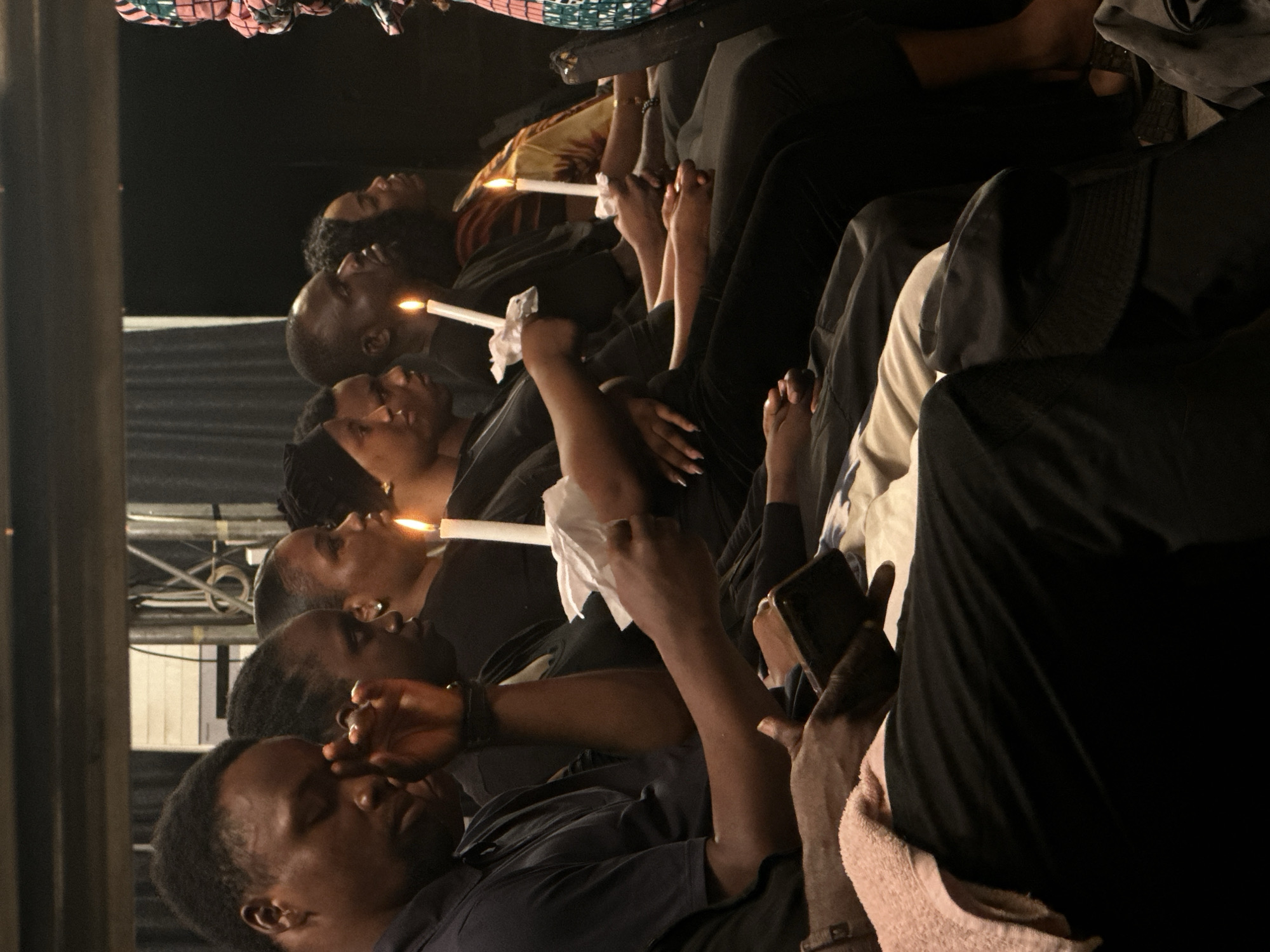 The image shows a group of Black individuals sitting in what appears to be an indoor setting, possibly a church or community hall in Ashipa, Nigeria. Most individuals are holding lit white candles. The lighting is dim, with the primary illumination coming from the candles and some overhead fixtures. The people are seated in rows, and their expressions suggest a somber or reflective mood. One person in the foreground is wiping their eye, and another is holding a tissue. There are various clothing styles visible, predominantly dark colors. The background is indistinct, with dark curtains or walls. The overall atmosphere is one of a memorial service or vigil.