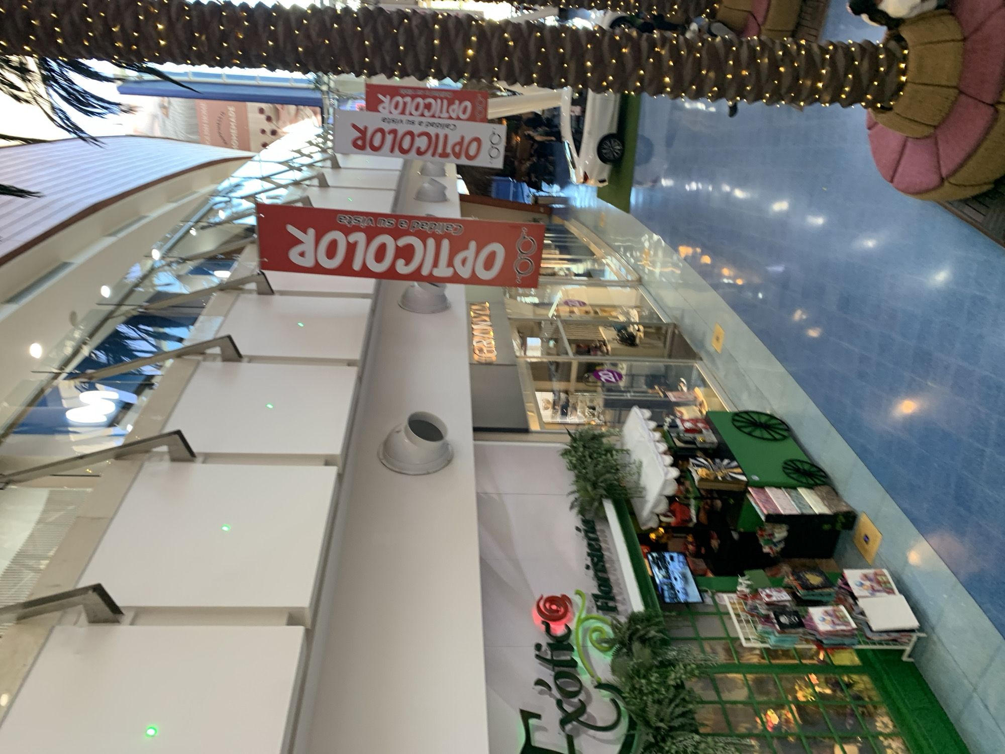 The image shows an indoor mall interior, possibly in Porlamar, Venezuela, with a wide, blue-tiled floor reflecting overhead lights and storefronts. On the left, a florist shop named "Exotic Floristeria" is visible, adorned with green foliage and displaying flowers and gift bags. A small green cart with wheels, resembling a flower stand, is situated in front of the shop. Further down the corridor, several red banners with white text reading "OPTICOLOR" are hung vertically. These banners suggest the presence of an optical store. In the background, a white car is parked on a green artificial turf area, and the upper levels of the mall show architectural features and more storefronts. Palm trees decorated with small lights are also present, adding to the ambiance. Several people are faintly visible in the background, walking or browsing. The overall scene depicts a well-lit, commercial environment.
