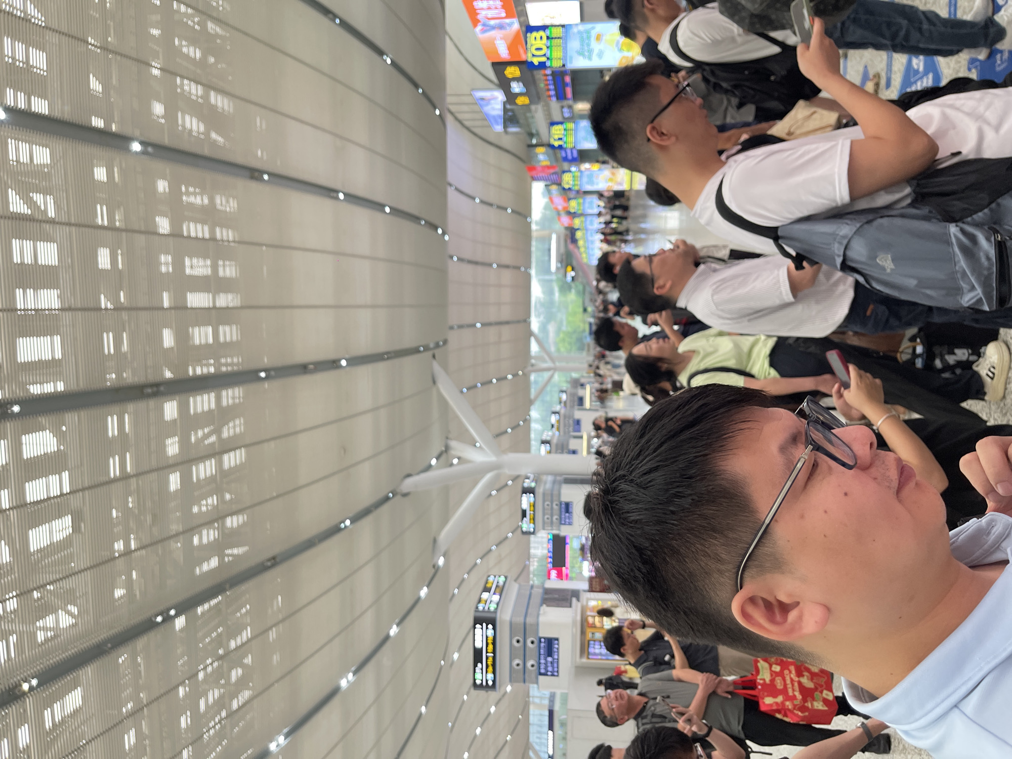 The image captures a crowded scene inside what appears to be a modern transportation hub, likely a train station or airport, in Longhua District, China. The ceiling is high and modern, with a patterned metallic structure and recessed lighting. A large crowd of people, predominantly adults, are seen standing and walking in various directions. Many are carrying luggage, such as backpacks and bags. People are dressed in casual attire. Some individuals are looking at their mobile phones. In the background, numerous digital display boards with colorful advertisements and information are visible, indicating a commercial and informational environment. The architecture suggests a contemporary public space designed to accommodate a large volume of travelers. The overall impression is one of a busy and active transit point.