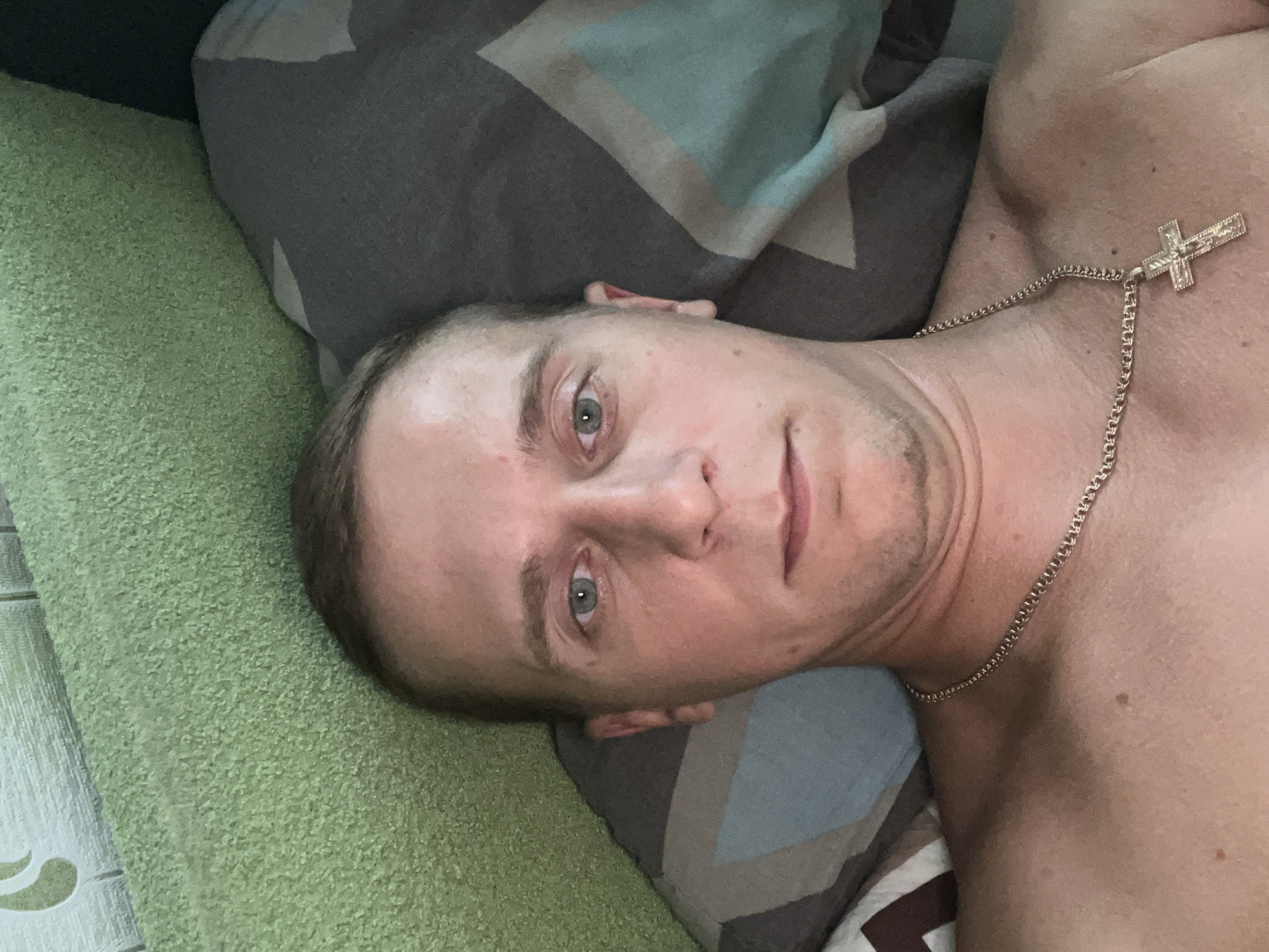 A man with light skin, light brown hair, and light blue eyes is lying on a bed, looking at the camera. He is wearing a gold chain with a cross pendant. The bed has pillows with a patterned fabric. The environment appears to be a bedroom, possibly in an apartment. The image quality is adequate for identification. The man's facial expression is neutral. No specific location within Minsk, Belarus can be determined from the image.