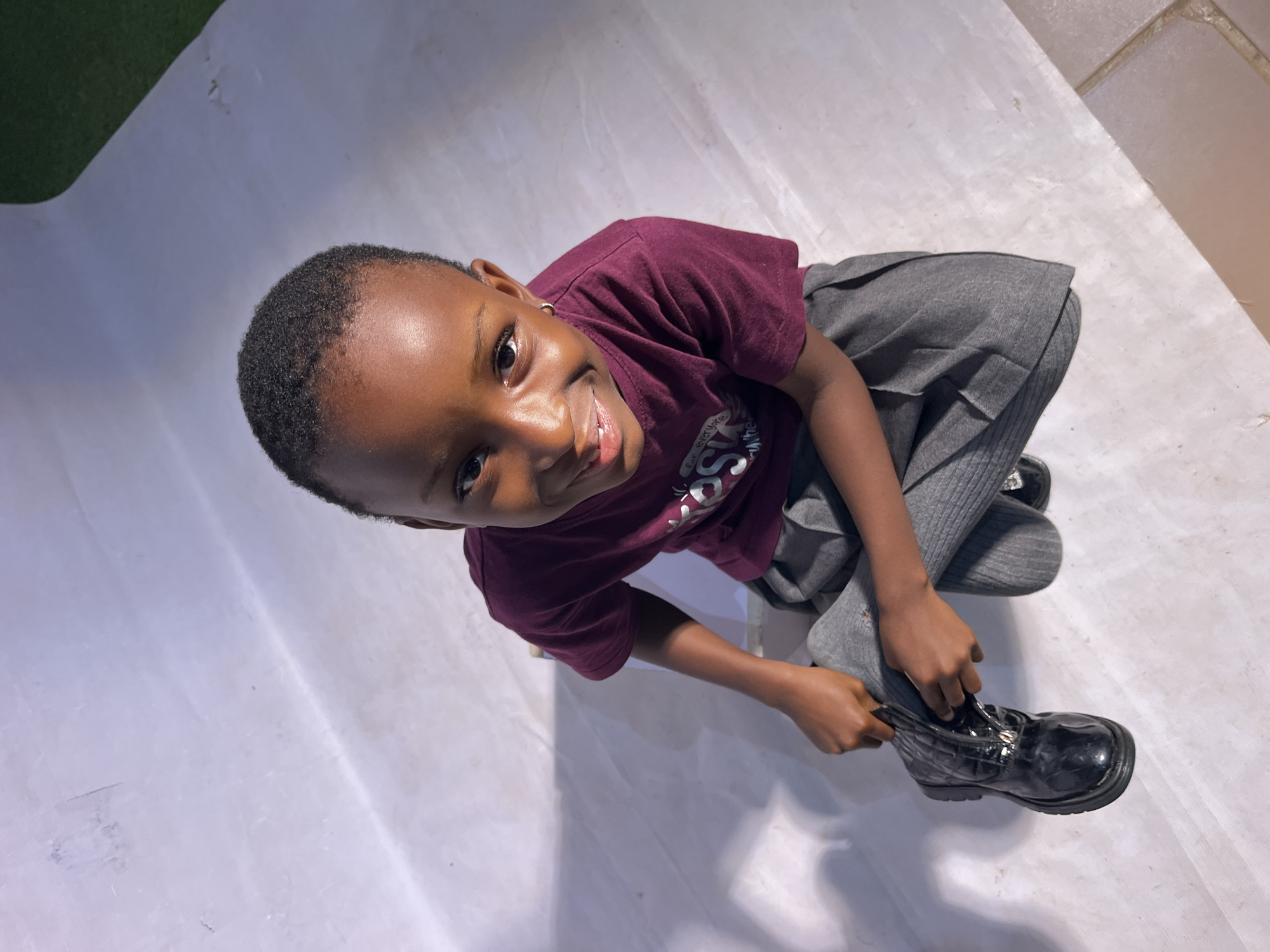 The image shows a young child, viewed from a high angle, in an indoor setting. The child, who appears to be a girl, is wearing a maroon t-shirt and a grey pleated skirt. She is in the process of putting on a black shiny shoe. Her face is turned towards the camera, and she has a bright, engaging smile. Her hair is short and dark. The background is predominantly white, with some areas of shadow, suggesting artificial lighting. There's also a hint of green and brown in the upper left and upper right corners, which could indicate elements of the environment beyond the main backdrop. The focus is on the child and her activity, capturing a moment of childhood engagement. No text is clearly visible in the image. The overall mood is cheerful and candid.