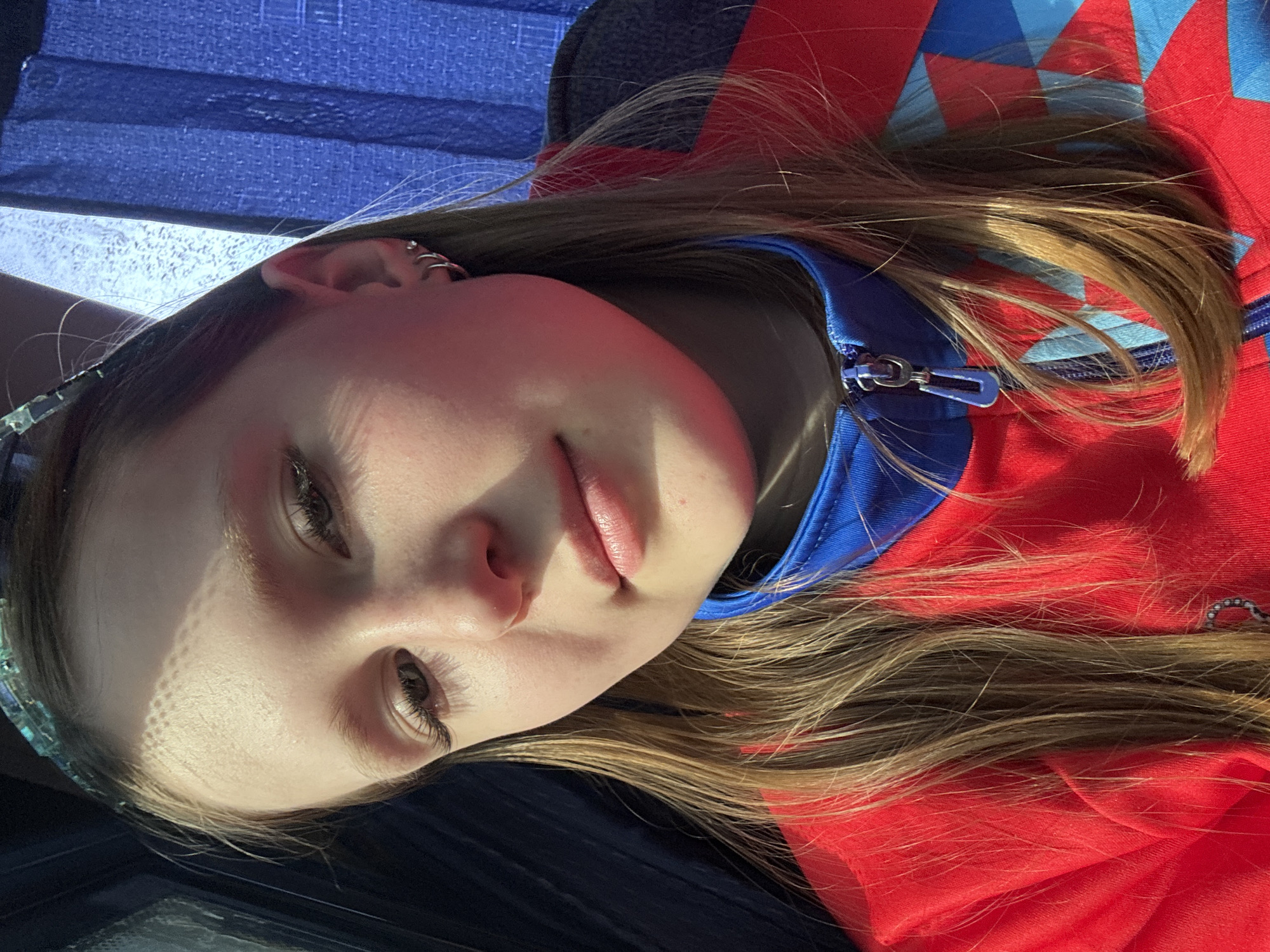 A young woman with long brown hair is lying down. She is wearing a red and blue zip-up jacket with the zipper partially undone. The background includes a dark blue material. It's not possible to know if the location is in Пальники, Russia.