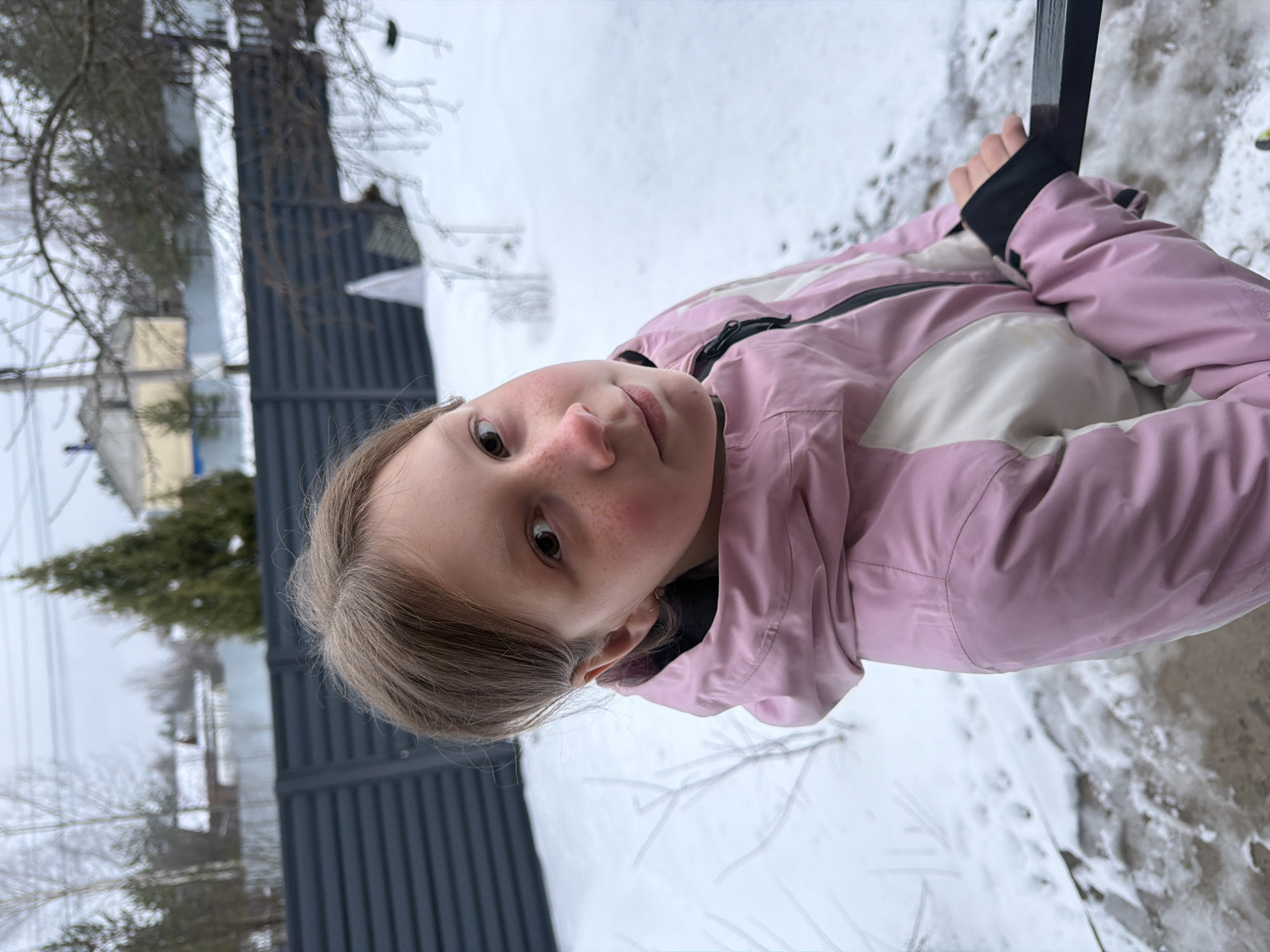 A young girl with fair skin, light brown hair, and freckles on her face is centered in the frame. She is wearing a pink and white jacket with a black zipper and dark-colored cuffs, holding onto a dark vertical bar. The background shows a winter scene in Markovo, Russia, with a snow-covered ground. A dark gray fence runs along the left side of the image, and a green evergreen tree is visible behind it. In the distance, there are glimpses of buildings and other trees, further suggesting an outdoor environment.