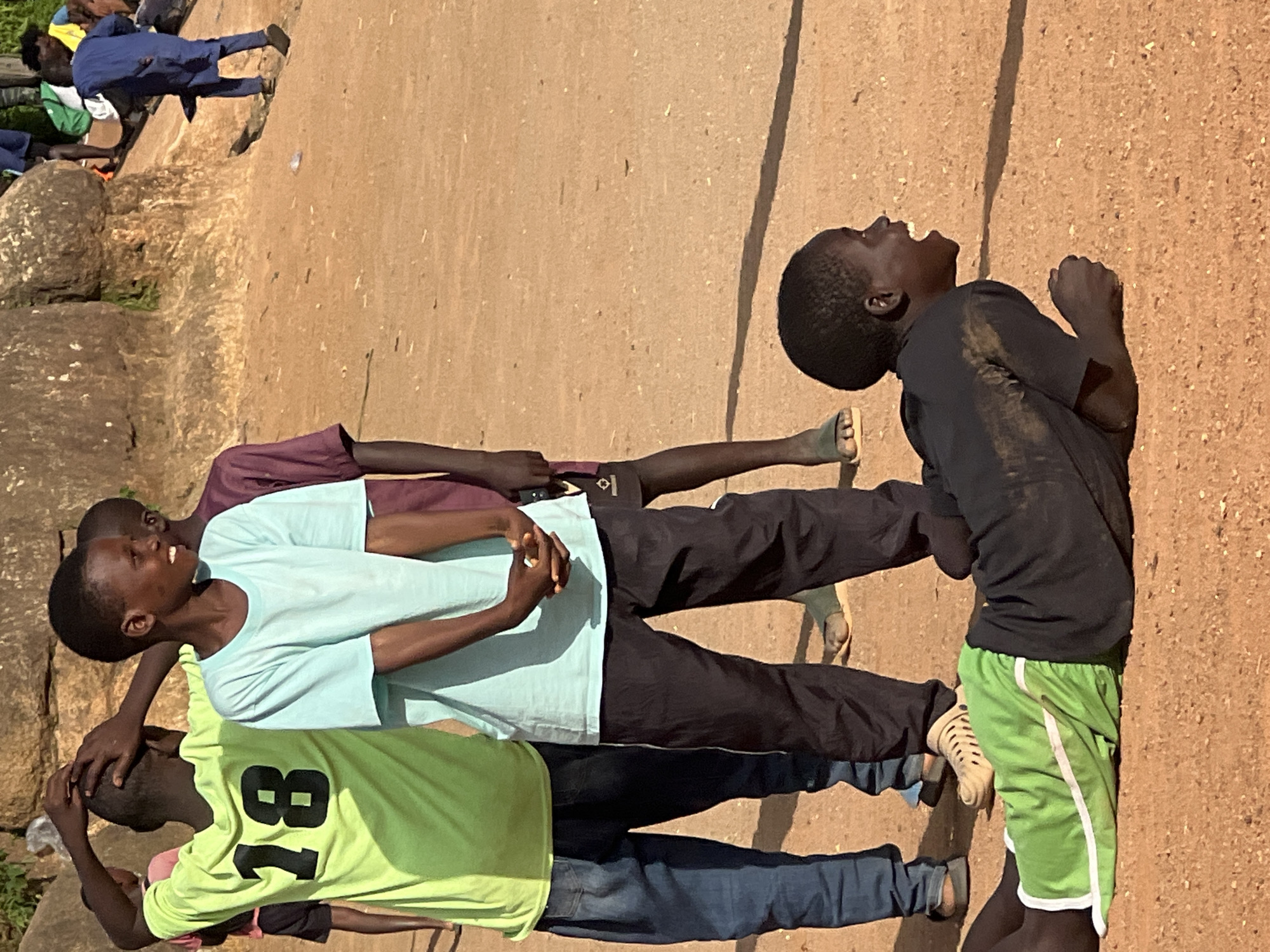 In the city of Jos, Nigeria, a group of young boys are gathered in an outdoor, sandy area with large rocks in the background. In the foreground, one boy lies on his stomach, wearing a black t-shirt and bright green shorts with a white stripe, his mouth open in an expression of surprise or exertion. Behind him, a boy stands with his hands clasped behind his back, wearing a light blue t-shirt and dark pants. To his left, another boy in a lime green t-shirt with the number "18" on the back, also wears dark pants. Further back, other boys are visible, one in a purple shirt and another in a blue traditional garment, suggesting a communal activity or gathering. The ground is predominantly sandy, with some sparse vegetation visible at the edge of the frame. The overall impression is a snapshot of youthful energy and interaction in a natural, informal setting.