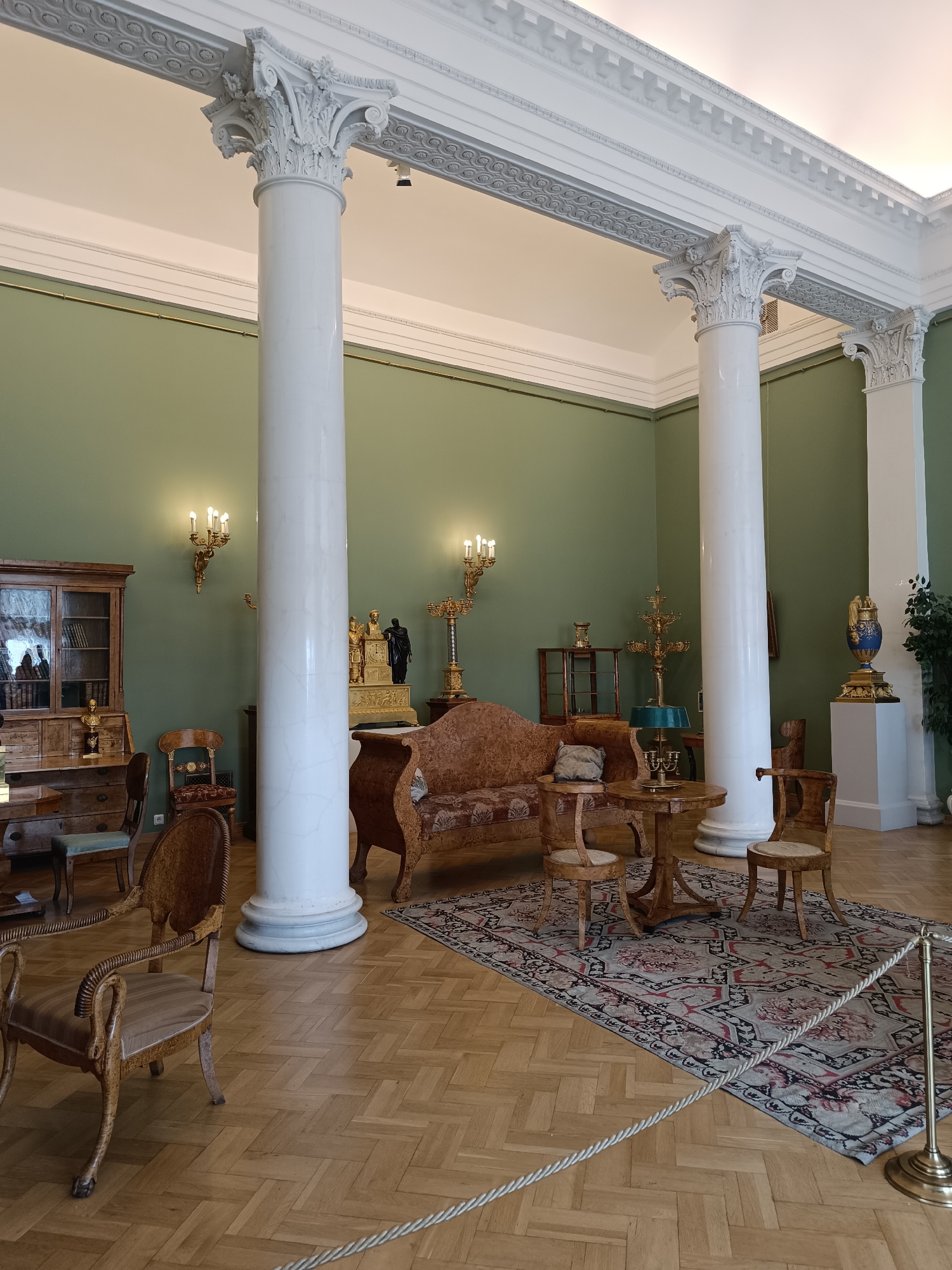 The image shows an interior view of a room with ornate columns and antique furniture. The room has pale green walls and a wooden parquet floor. In the foreground, there are several wooden chairs and a small round table. A large rug with a floral pattern is placed in front of a patterned sofa. Behind the sofa, a large clock with figurines is visible. To the left, a wooden cabinet with glass doors displays books. Two sconces with multiple lights are mounted on the wall. In the background, another column supports the ceiling. This image likely depicts a room in the Hermitage Museum in Saint Petersburg, Russia, given the architectural style and the presence of historical artifacts and furnishings. There are no people visible in the image.