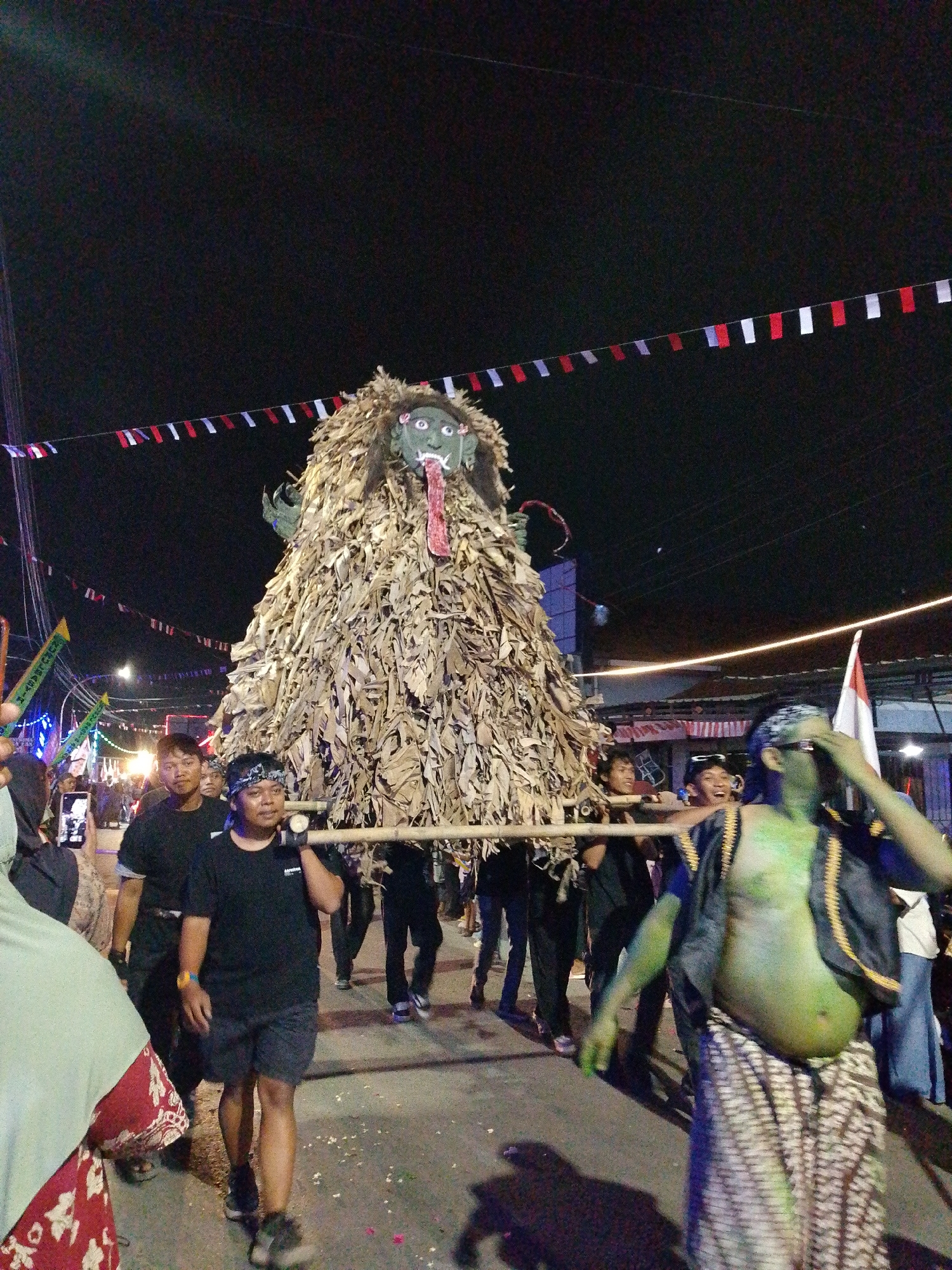 This photograph captures a nighttime procession in Pagongan, Indonesia, featuring a large, effigy-like structure resembling a monstrous figure. The effigy is covered in dried leaves and has a grotesque green face with a protruding red tongue. It is being carried on bamboo poles by several men walking in the street. In the foreground, a man with green-painted skin and a patterned sarong is walking towards the camera. Other individuals are visible in the background, some holding flags and banners, and decorative lights illuminate the scene. The setting appears to be a street lined with buildings and utility poles, with strings of small flags strung across the sky. The atmosphere suggests a cultural or religious event.