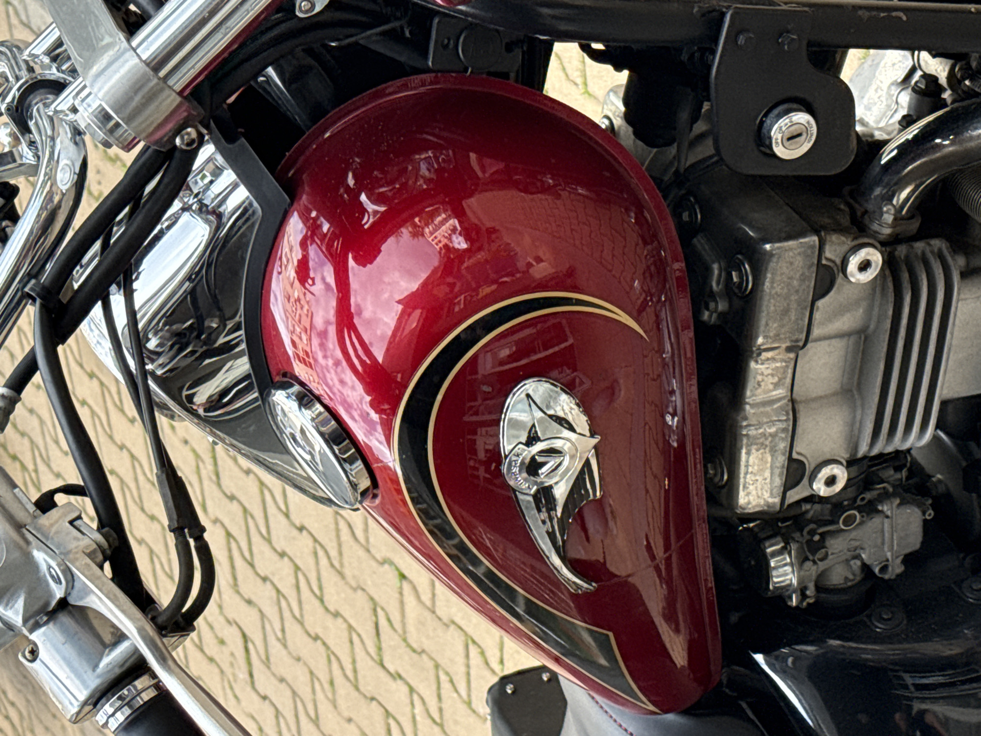 The image displays a detailed, close-up view of the right side of a motorcycle, primarily focusing on its fuel tank, handlebars, and engine components. The motorcycle features a glossy, deep red fuel tank with black and gold pinstriping, and a chrome emblem on its side that incorporates a stylized winged design and the word "Kawasaki." Above the tank, chrome handlebars, brake/clutch levers, and various cables and hoses are visible. Below the tank, metallic grey engine components, including cooling fins and part of a carburetor, are present. An ignition switch, labeled "OFF," "ON," and "IGNITION," is mounted on a black bracket to the right of the engine. A portion of a black motorcycle seat with red stitching is visible on the far left. The highly reflective surfaces of the tank and chrome elements show distorted reflections of an outdoor environment, including what appears to be buildings, trees, and sky. The ground visible in the background is a paved surface composed of light-colored, rectangular blocks. There is no specific visual information within the image to confirm its location in Sofia, Bulgaria.