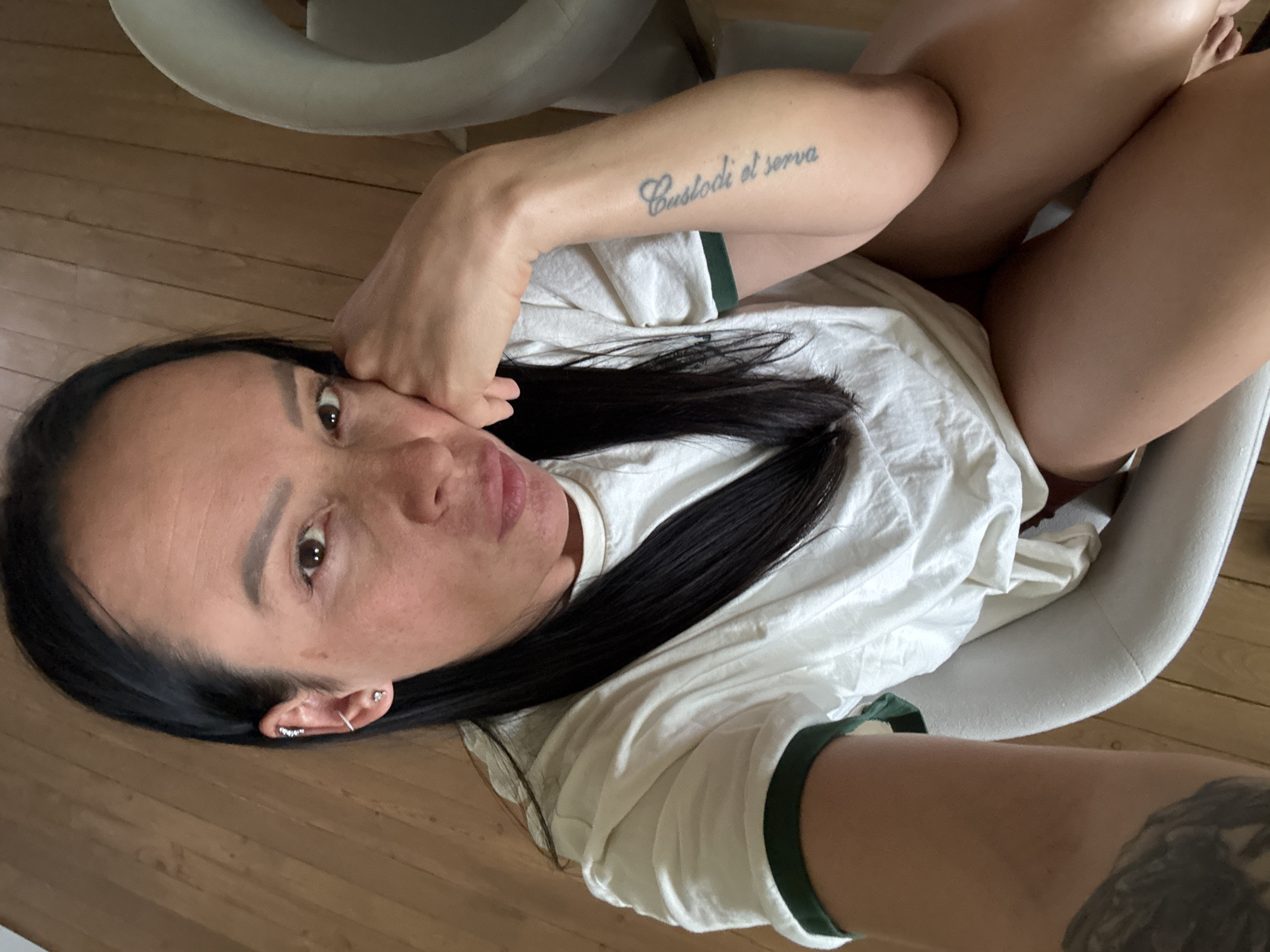 A woman with long, dark hair is lying in a light-colored chair. She is wearing a white t-shirt with green trim. There is a tattoo on her left arm that reads "Custodi et serva." There is a tattoo on her right arm as well. The background is a wooden floor. The location of this image cannot be identified, and there is nothing to suggest it is in Chaikovsky, Russia.