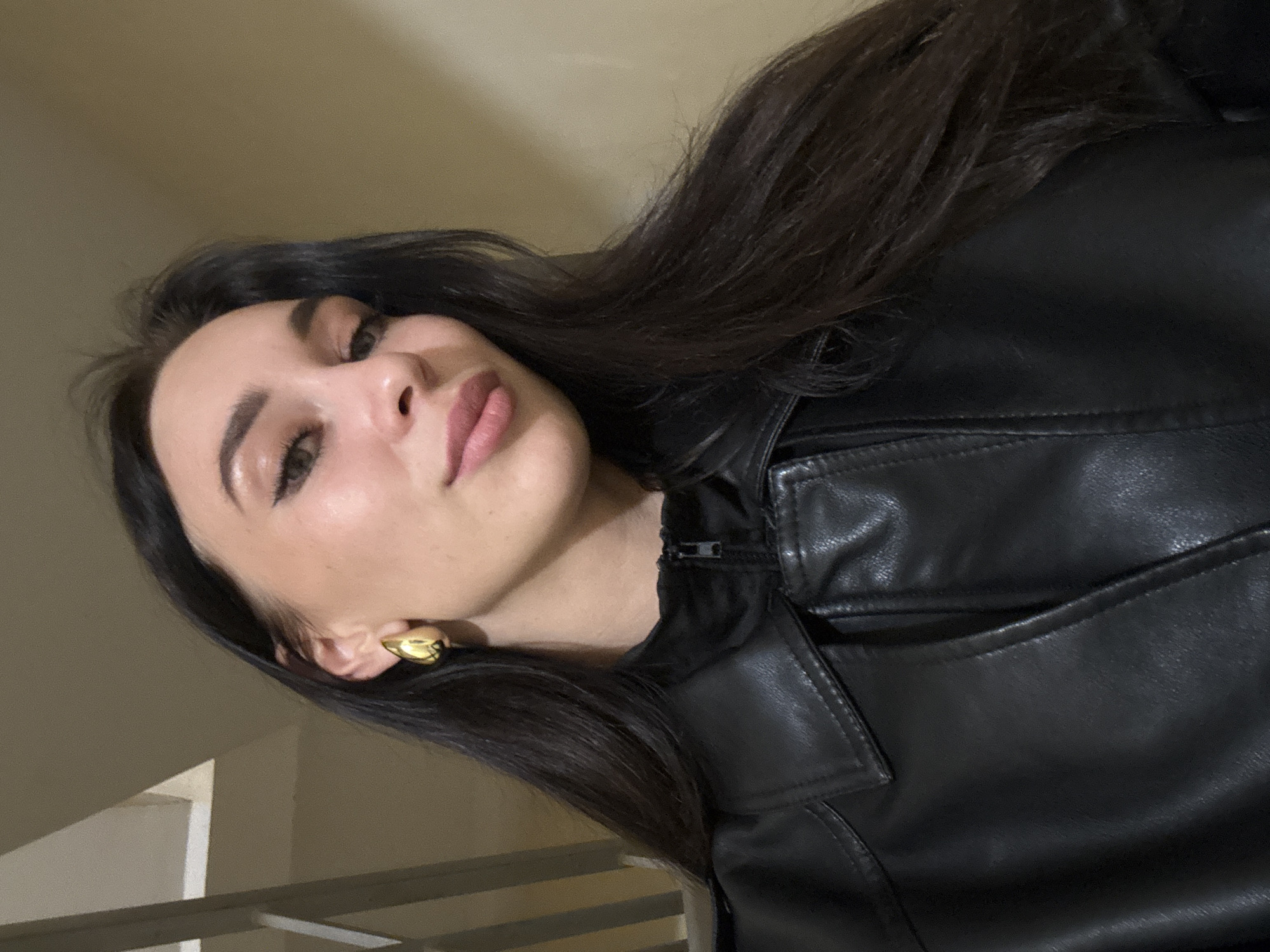 A young woman with olive skin, dark brown hair, and light brown eyes is wearing a black leather jacket and gold earrings. She is positioned at an angle in the frame, looking towards the viewer. The background appears to be indoors, with part of a light-colored wall and a metal railing visible. The setting's exact location within Chaikovsky, Russia, is not discernible from the image.
