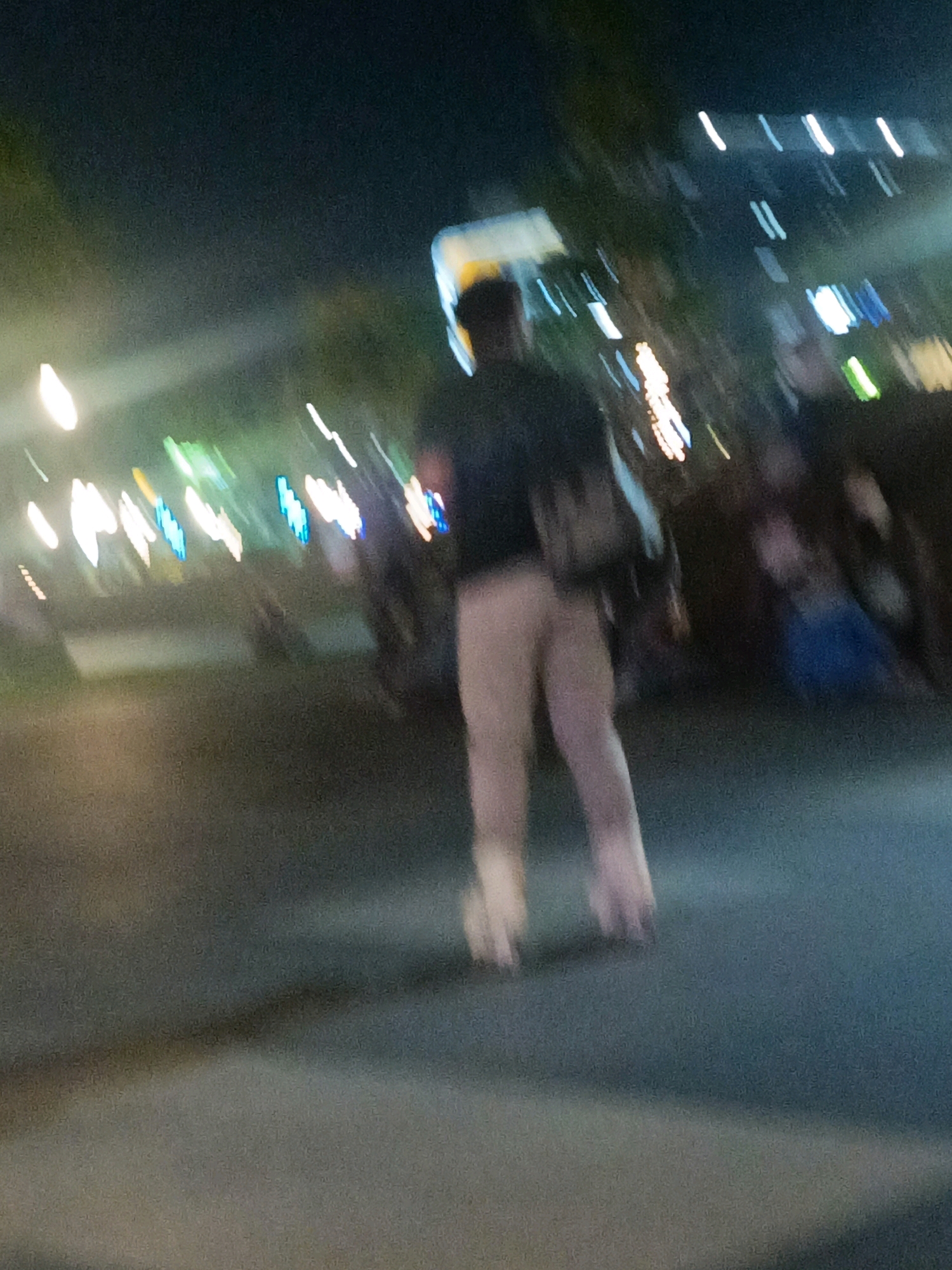 An out-of-focus, nighttime image shows a person wearing light-colored pants and a dark top, possibly rollerblading or roller skating on a paved surface in the city of Sidoarjo, Indonesia. The background is blurred, featuring streaks of colored lights that suggest a vibrant urban environment, possibly a street or a promenade. The lighting is predominantly artificial, with bright white and blue lights creating a dynamic, streaky effect due to the motion. The overall scene conveys a sense of movement and urban nightlife.