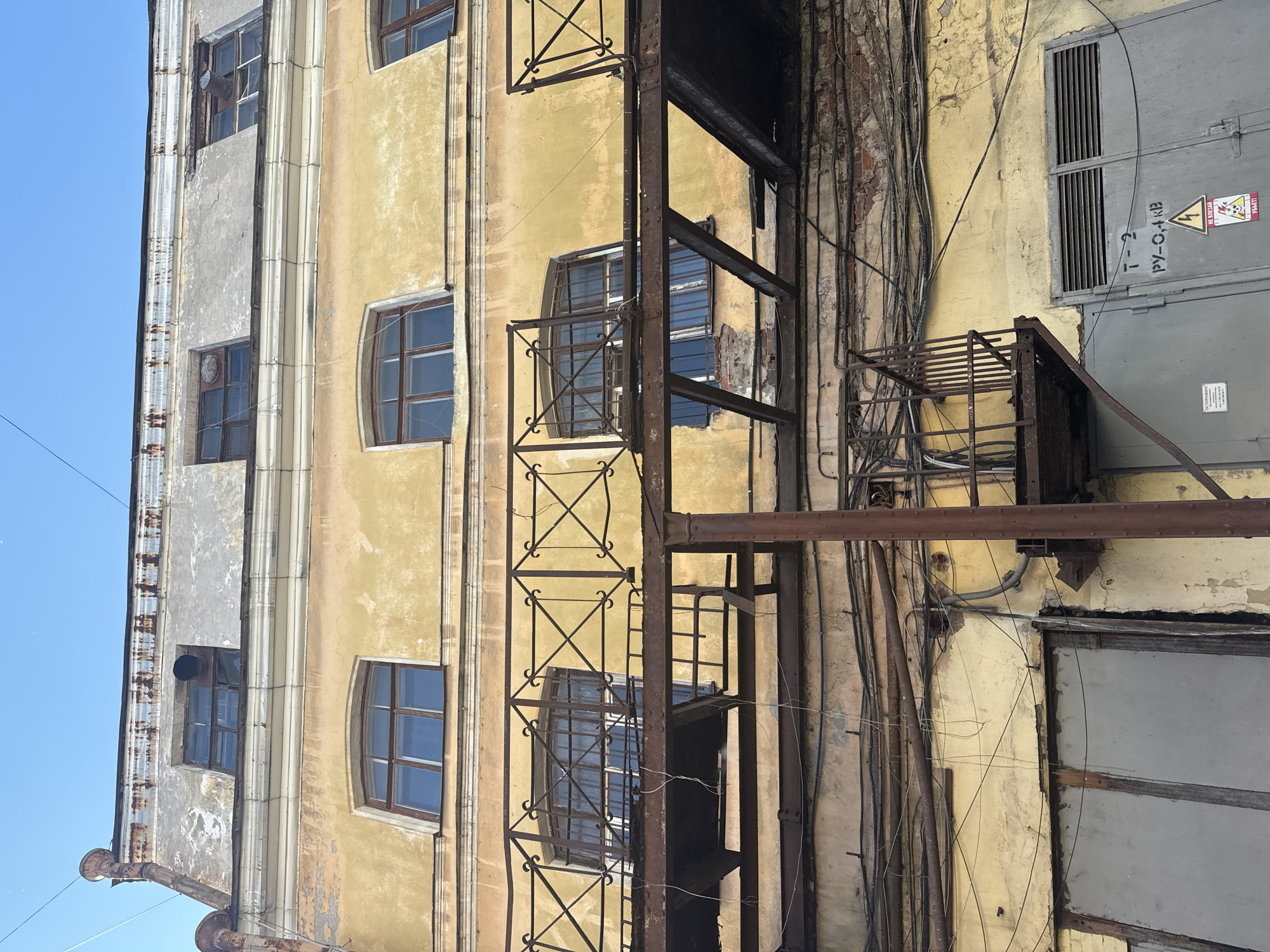 The image shows the exterior of an old, weathered building in Saint Petersburg, Russia. The building's facade is painted in a faded yellow, with areas of peeling paint and visible brickwork. A corrugated metal downspout runs along the left side of the building, showing signs of rust.
Several windows are visible, some with intricate wrought-iron grates. One window has a ventilation pipe protruding from it. A rusty metal fire escape or external staircase structure runs vertically up the building, with platforms and railings. Numerous electrical wires and conduits are haphazardly attached to the facade, adding to the building's industrial and aged aesthetic.
On the right side of the building, there is a large grey metal door or panel with louvers and a smaller panel below it. This area also features warning signs, including one with a lightning bolt symbol indicating danger, and another with text that appears to be in Cyrillic script. Some visible text on the grey panel reads "Т-2" and "РУ-С,4, КЗ". Another sign at the bottom has Cyrillic text as well.
The sky is a clear, bright blue, suggesting it is daytime. The overall impression is one of urban decay and a utilitarian, possibly industrial, setting. The light suggests a sunny day, but the weather conditions are not explicitly stated. No people are visible in the image. The scene evokes a sense of history and the passage of time, with the building bearing the marks of its age and use.