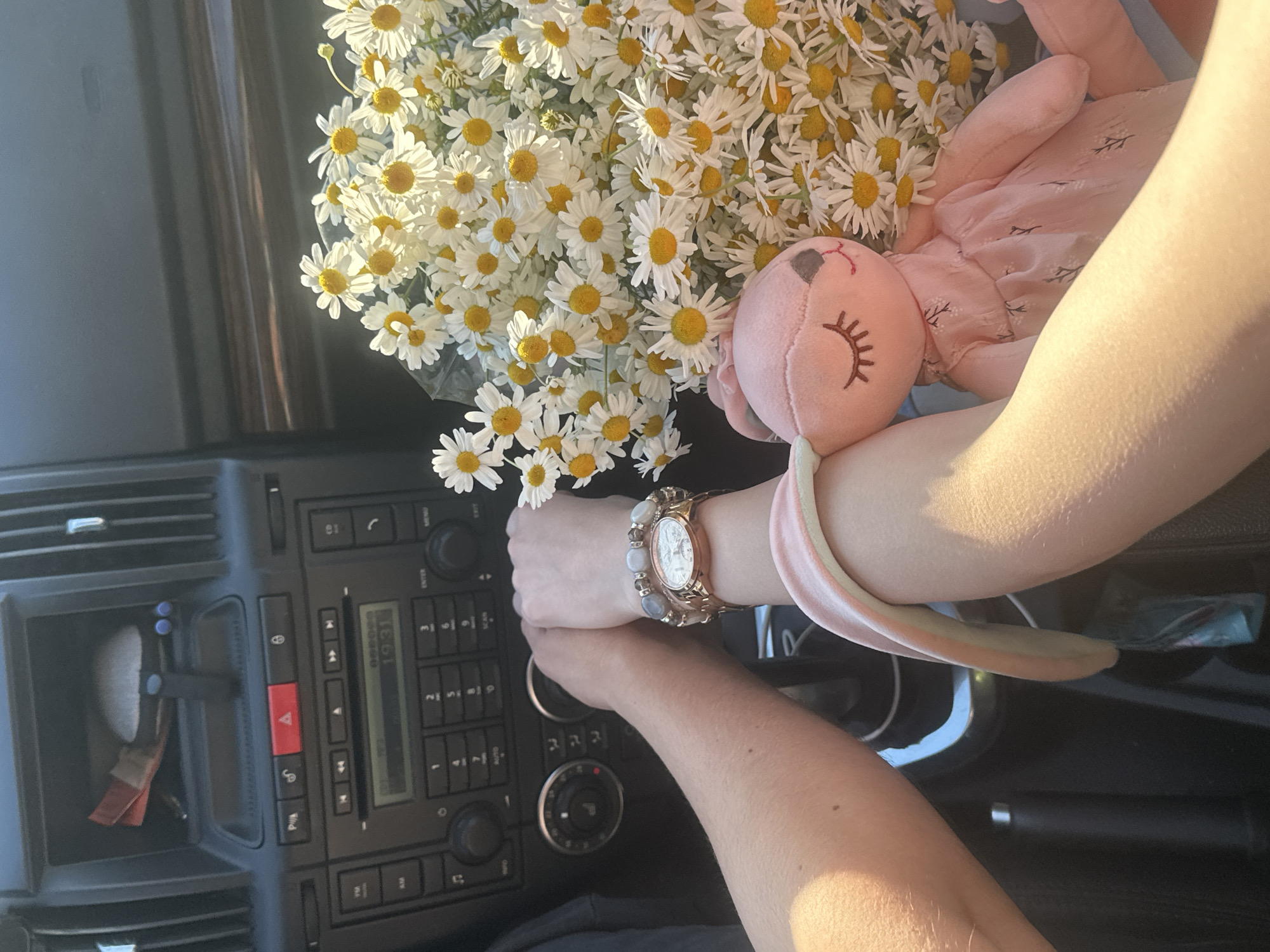 This media file features a close-up view of a person's arms and hands inside a vehicle. The setting appears to be the interior of a car, with visible dashboard controls and a storage compartment. A bouquet of white daisies with yellow centers is prominently displayed, along with a pink stuffed animal that resembles a bunny. The person is wearing a watch on their left wrist, which has a rose gold band and a white face with Roman numerals. A light pink and white headband or bracelet is also visible on the right arm.
The image captures a moment of stillness, possibly during a car ride or a pause in the journey. The lighting suggests it is daytime, with sunlight casting a warm glow on the scene, indicating a pleasant weather condition. The focus on the flowers and the stuffed animal suggests a theme of nature, comfort, or perhaps a gift. The visible dashboard details include buttons, knobs, and a display screen showing "19:31", which could indicate the time. The storage compartment contains a few items, including what appears to be a pen and some red and white wrappers. The overall mood conveyed is peaceful and perhaps sentimental. The location context of Khimki, Russia, provides geographical information but doesn't offer specific visual cues within the image itself. There is no discernible text other than the time displayed on the car's screen.