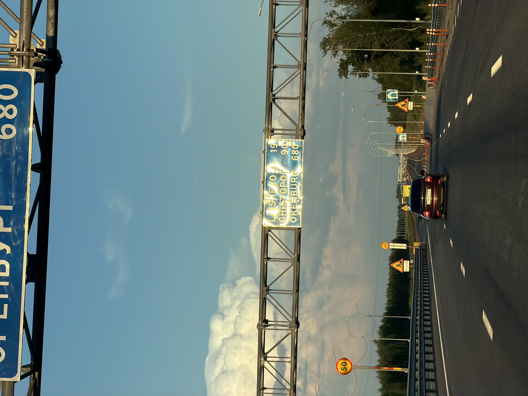 The image depicts a highway scene in Verkhnie Karmachi, Russia, likely during the daytime given the bright sky. The perspective is from a vehicle on the road, looking forward. The road is paved and has clear lane markings. To the left, a large blue signpost with white lettering is partially visible. The visible text on this sign is "680" and some illegible Cyrillic characters. Above the road, a metal framework supports another sign. This sign has yellow and blue sections and displays text in Cyrillic, including "190" and "680," along with "LAST CHISTOPO" and "ORENBURG."
Along the right side of the road, there are several traffic signs. One prominent yellow triangular sign shows a person walking. Another circular sign indicates a speed limit of "50." Traffic cones are also visible along the roadside, suggesting possible road work or a temporary lane closure. A dark-colored car is driving ahead on the highway. The overall scene is one of travel on a rural or suburban highway, with typical road infrastructure and signage. The sky is blue with some white clouds, indicating fair weather.