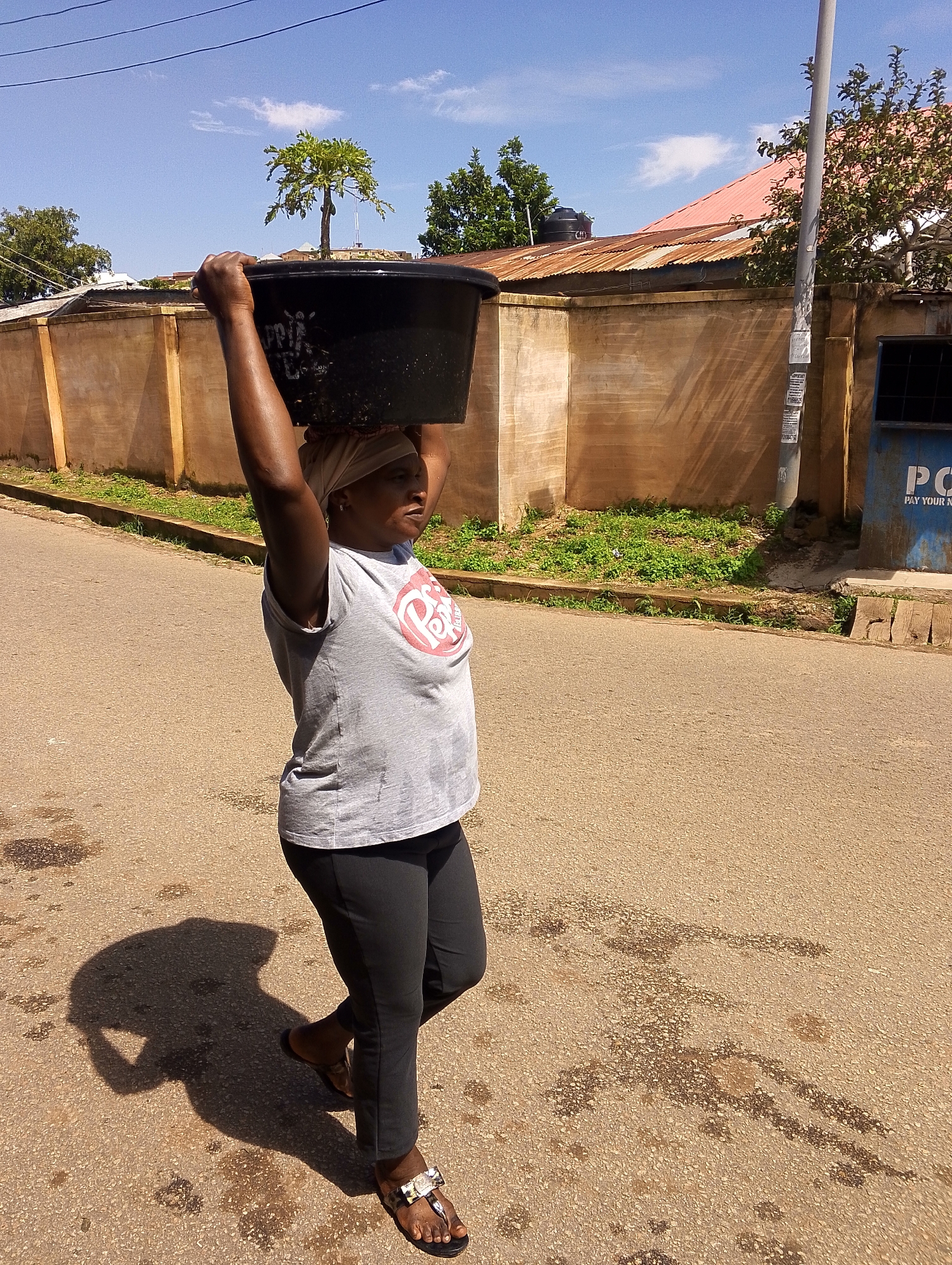 A woman walks along a paved street carrying a large black tub of water on her head. She is wearing a grey t-shirt with a red logo and dark pants. Her shadow is cast on the street behind her. A wall with some grass and trees lines the street. A building with a red roof is visible in the background. A blue dumpster with "PO" written on it is also in the background.