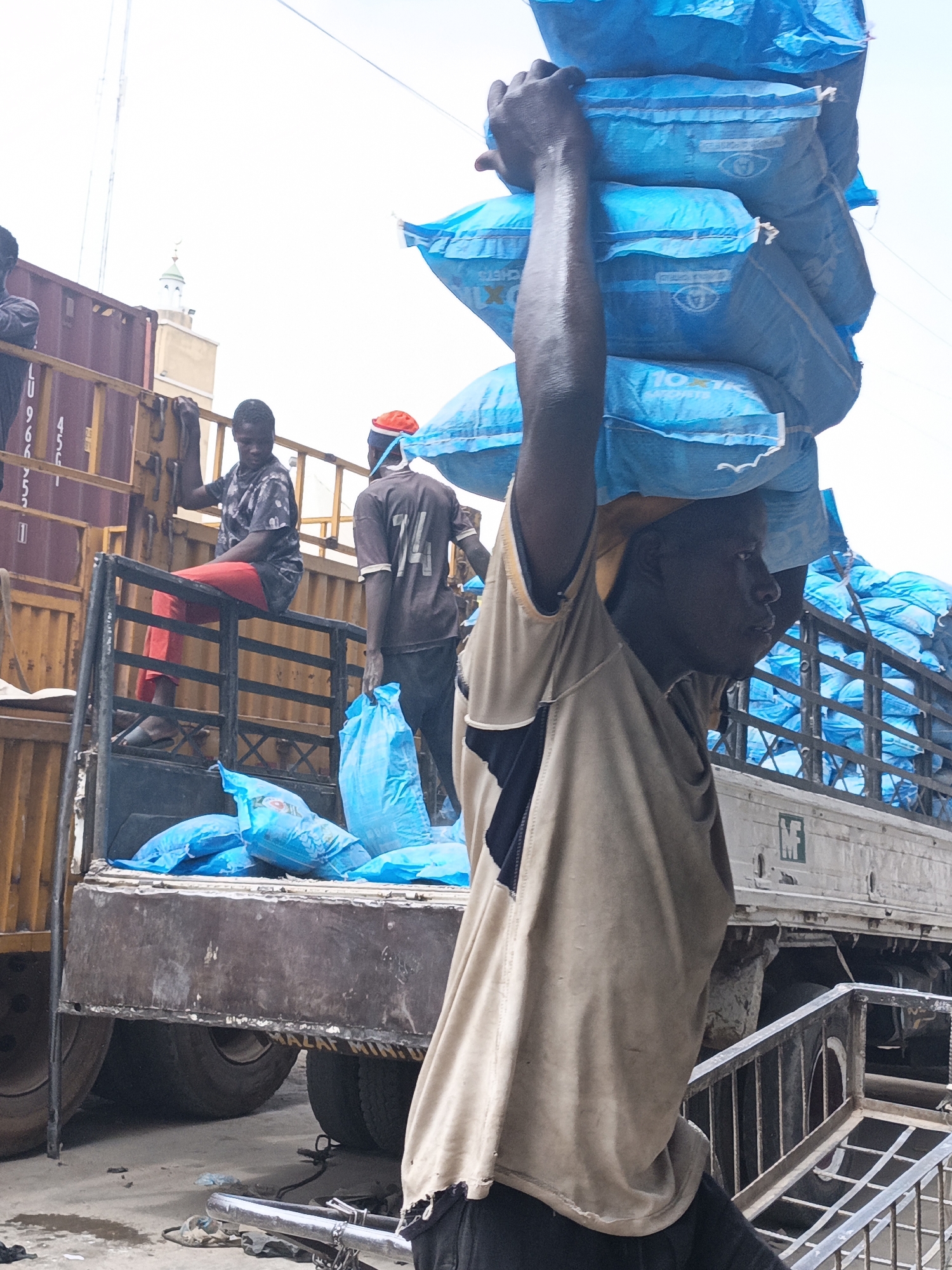 In Fagge C, Nigeria, a man carries a stack of blue bags on his head. He wears a tan shirt with a black patch on the shoulder. In the background, a truck is partially visible, loaded with the same blue bags. Two people are on the truck, one sitting on the railing in red pants and the other standing. The truck is parked next to a shipping container. A building with a green and white tower can be seen in the background.