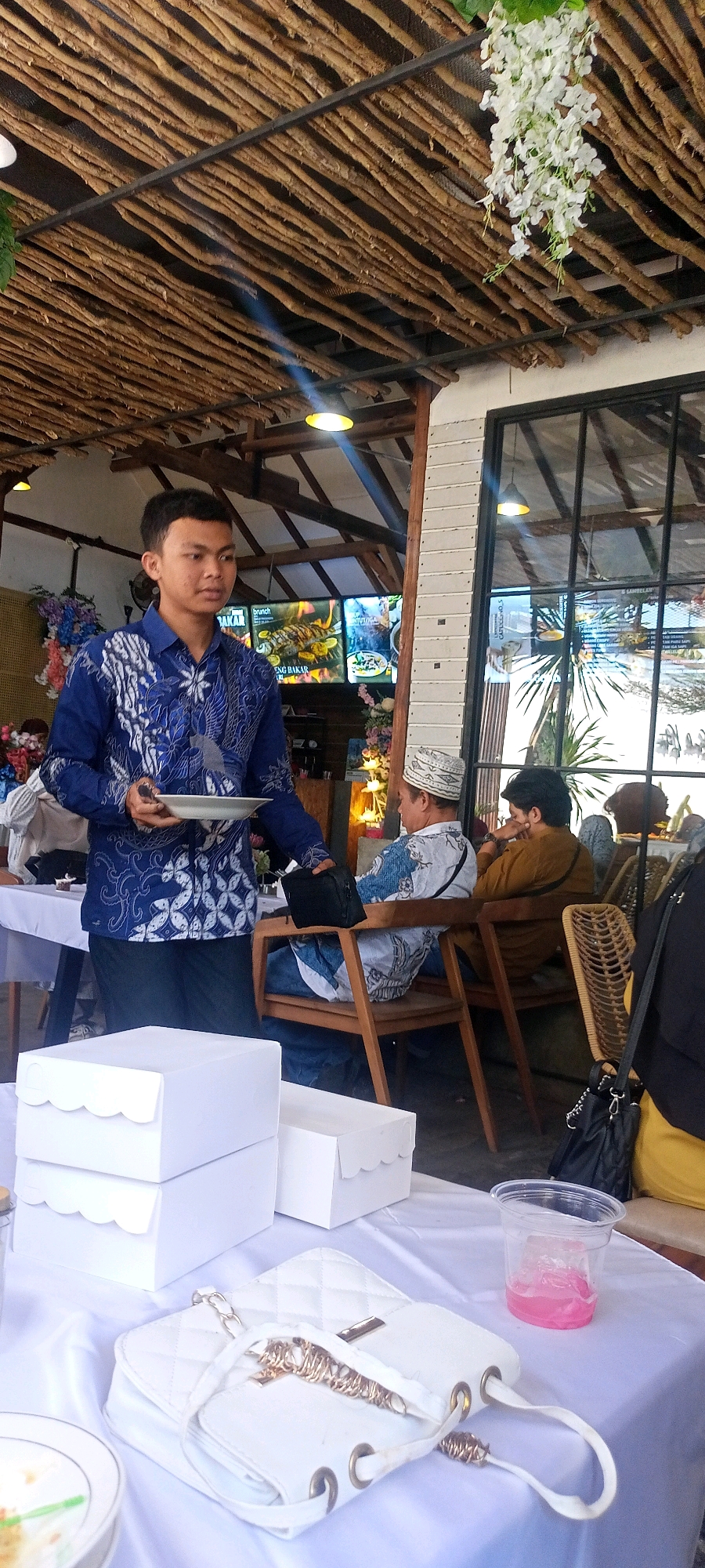 In Desa Ponokawan, Indonesia, at what appears to be an outdoor cafe or restaurant with a thatched roof, a young man in a blue batik shirt is serving food on a white plate. He is standing near a table laden with white cake boxes and a white quilted purse. In the background, two men are seated in chairs, one wearing a white cap and batik shirt, and the other in a brown shirt. A large window with black framing looks out onto a green outdoor space, with plants visible. Menu boards are displayed on the wall behind the young man, showing images of food, including grilled fish. The lighting suggests it is daytime, with some natural light filtering through the roof.