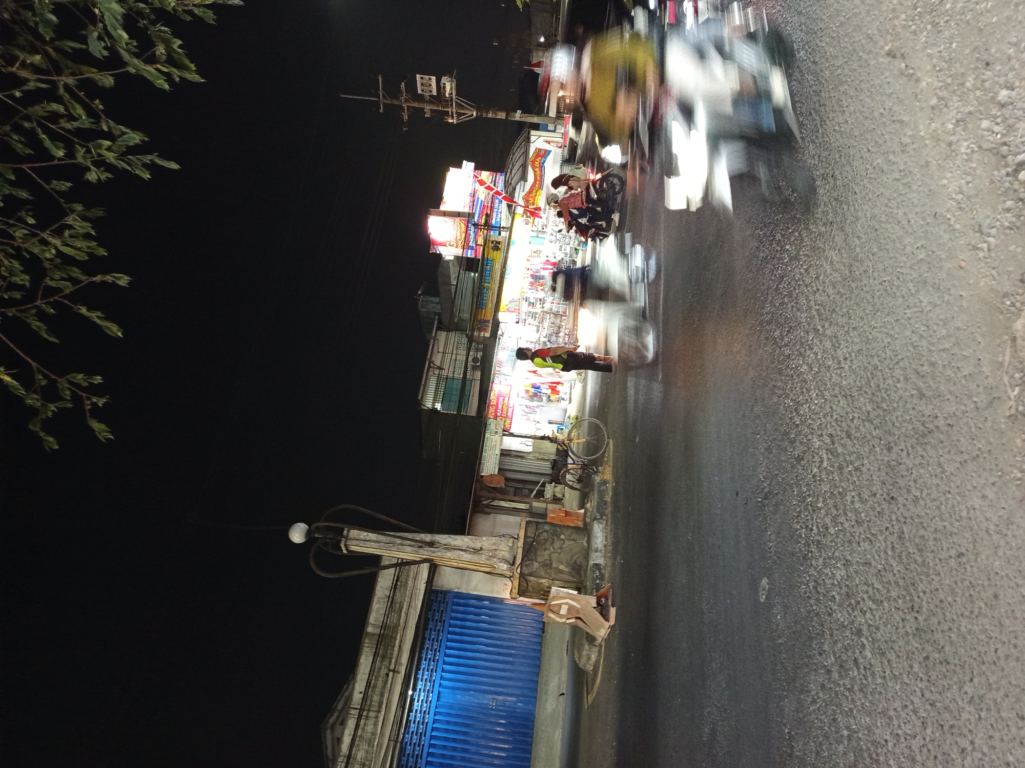 At night on a street in Buduran, Indonesia, traffic is moving with blurred motion. Several motorcycles are visible, with two riders on one and at least two other motorcycles with blurred riders in motion. A person in a yellow and green shirt stands on the sidewalk, facing away from the camera. In the background, there are buildings with illuminated signs. A blue roller door is prominent on one of the buildings. A bicycle with an orange seat is parked on the sidewalk next to a concrete structure topped with a light fixture. Traffic cones are placed on the side of the road. The sky is dark, with some tree branches visible in the upper right corner. The street surface is asphalt.