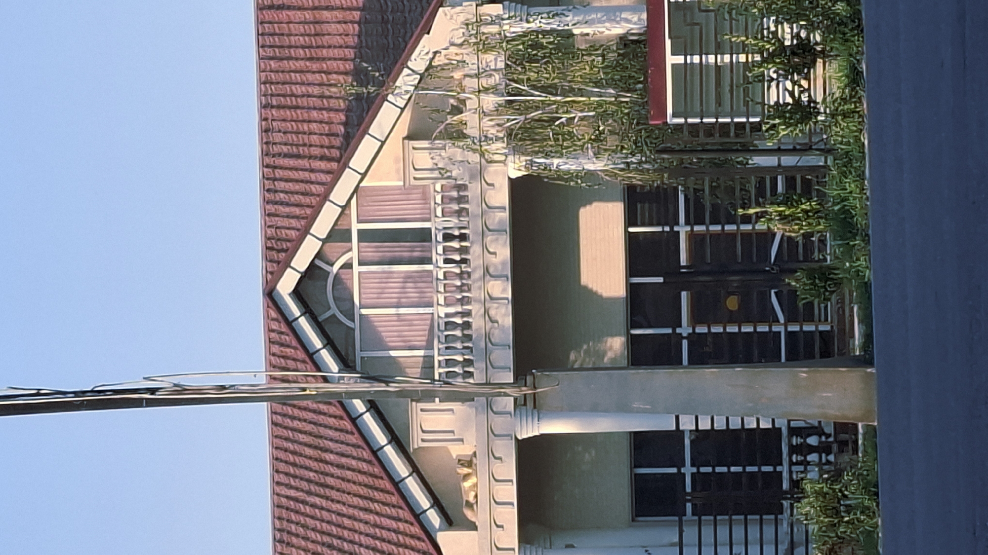 This image captures a residential building on a clear day in Fergana City, Uzbekistan. The building features a prominent tiled roof and a second-story balcony with white railings. A large, light-colored pillar stands in front of the structure. The ground floor has large windows or doors with metal grates. Lush green vegetation surrounds the base of the building. The sky above is a pale blue.